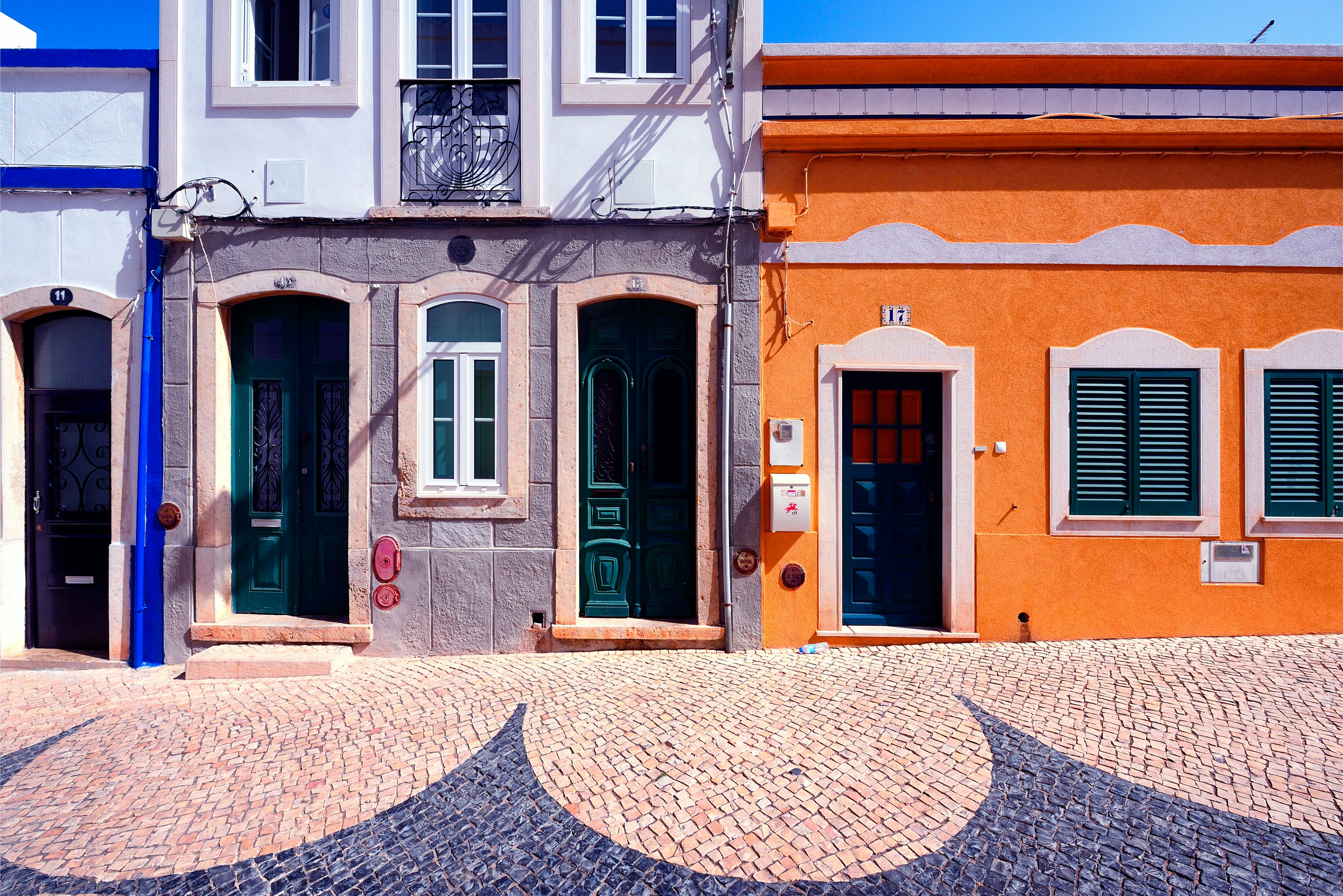 Black and beige cobbles form a curved pattern on the sidewalk of a street lined with houses painted in gray and orange.