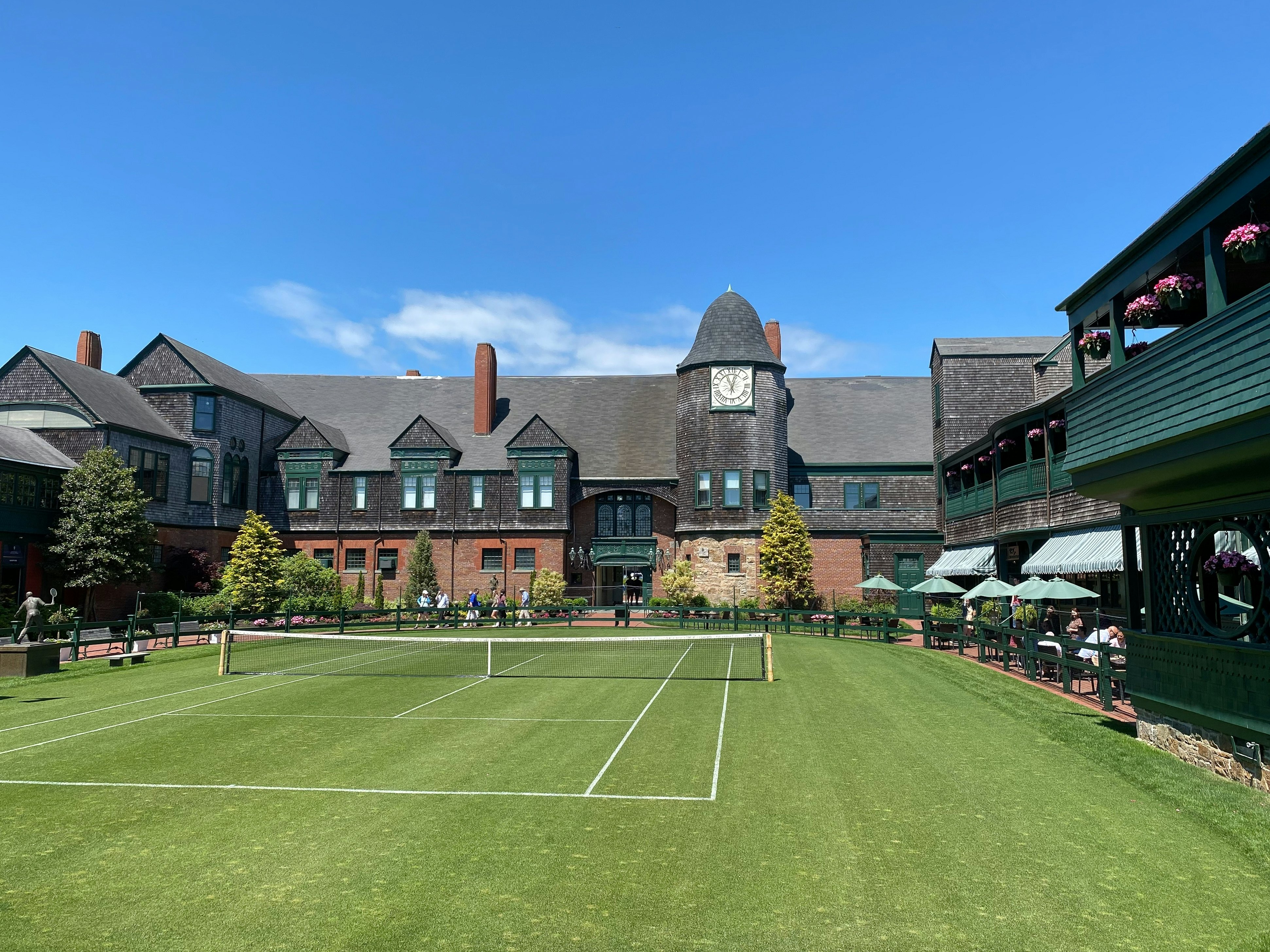 A grass tennis court surrounded on all sides by a brick and cedar-shingled building.