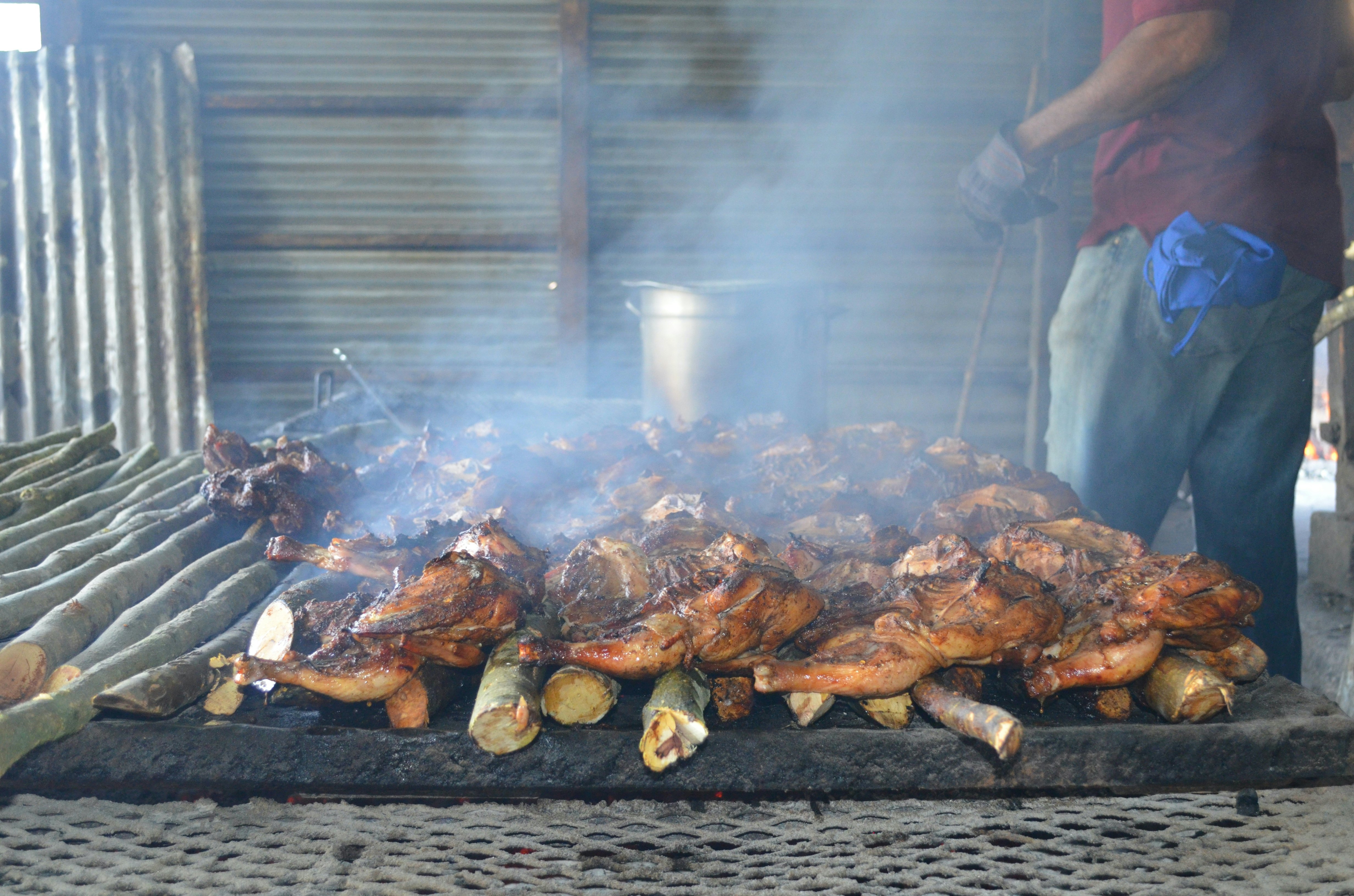 A chef stands near a charcoal fire with whole chickens on the grill and smoke rising above.