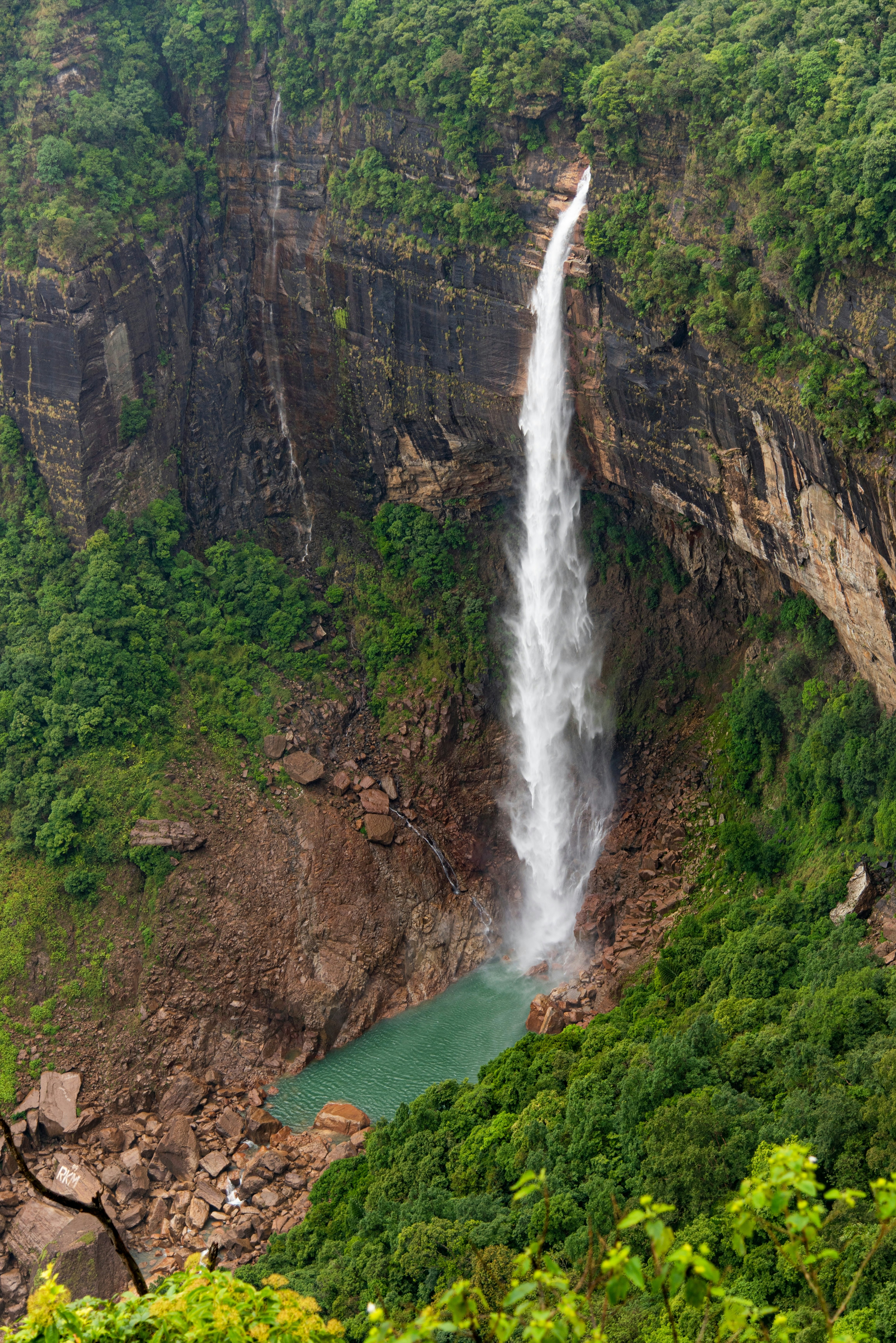A tall waterfall cascading over the edge of a cliff and plunging down into a turquoise pool of water among dense jungle.