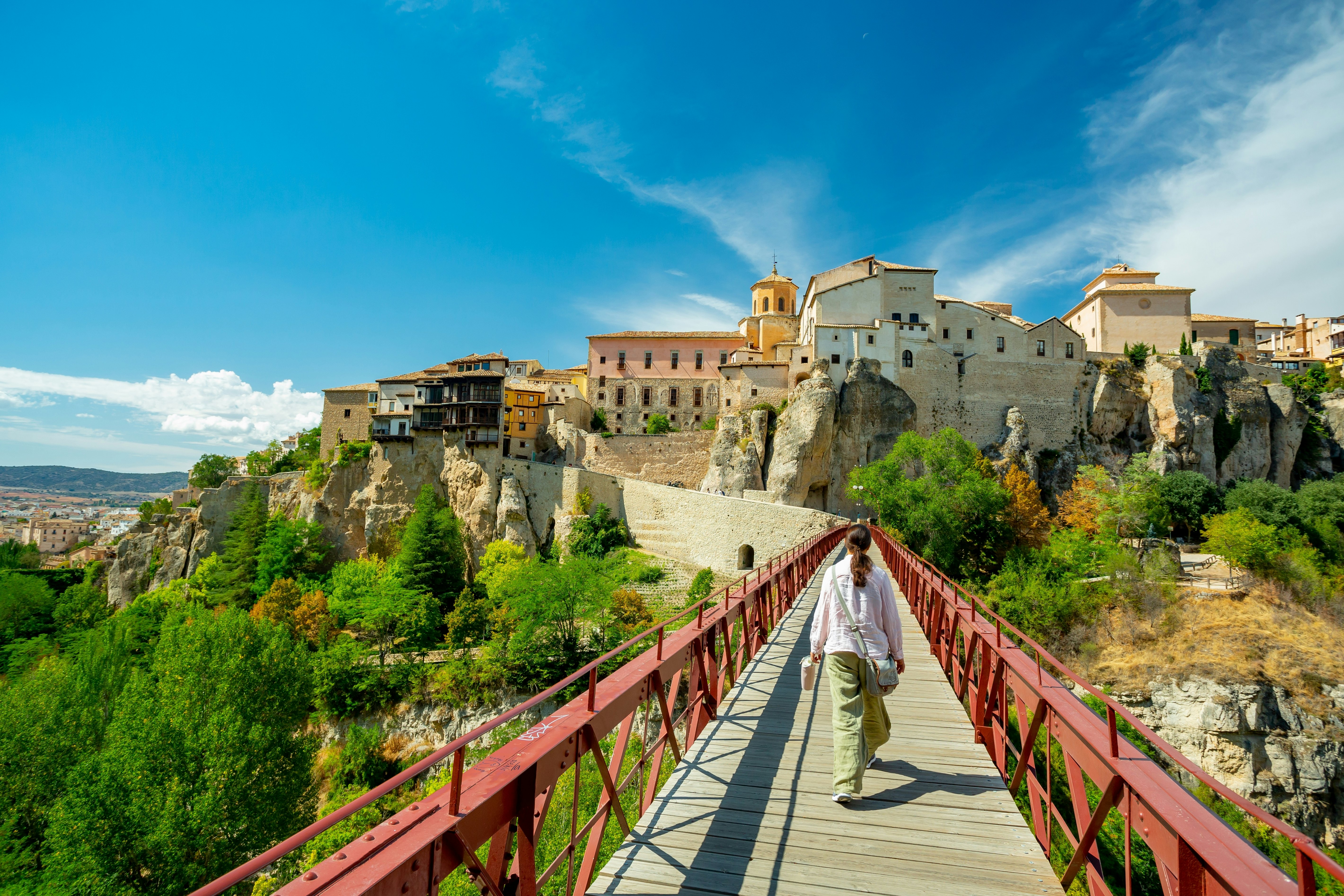 A woman crossing the San Pablo bridge with Cuenca's hanging houses ahead of her. Blue sky above.