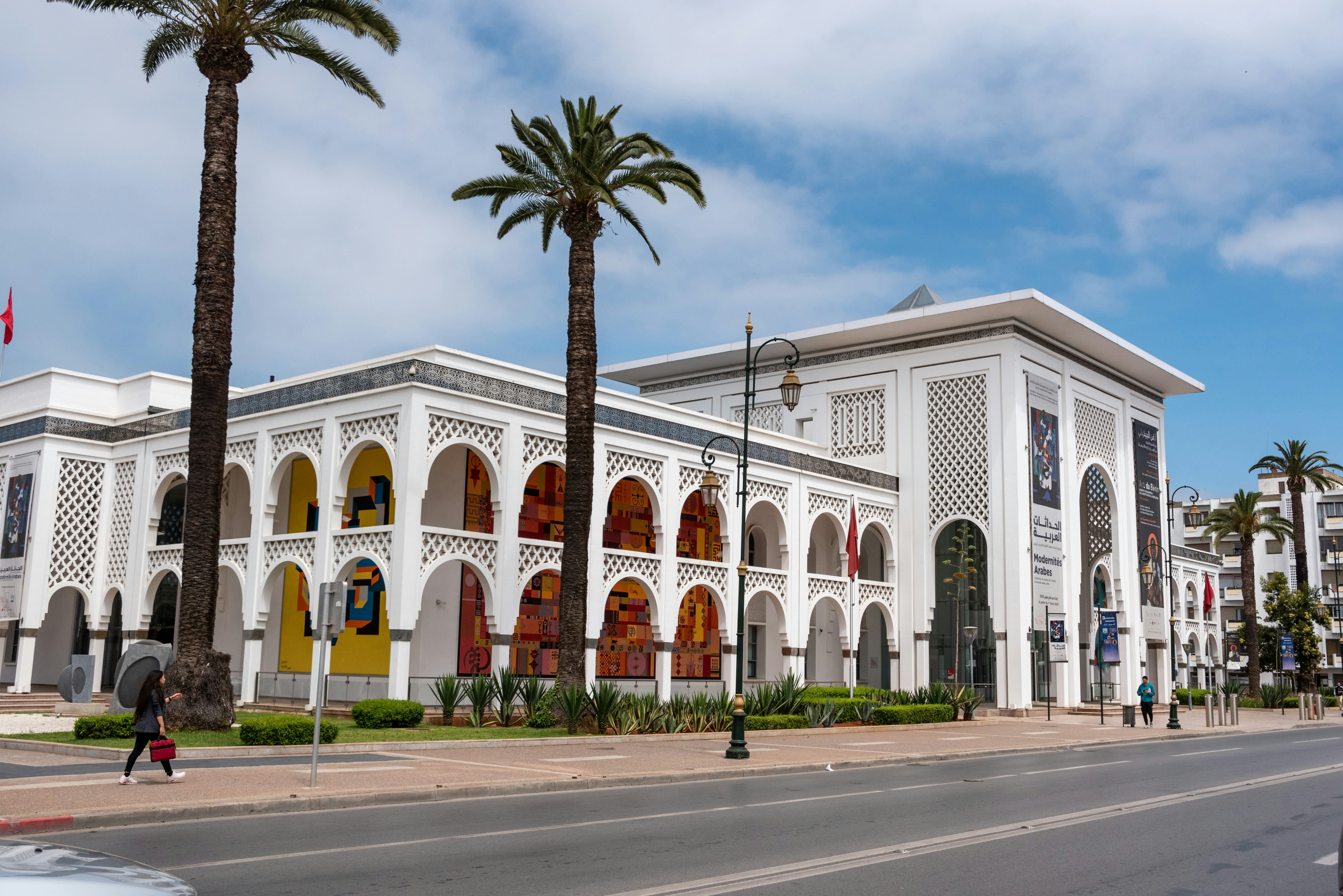 A sprawling white building with latticework and large arches with a few palm trees around it on a city block