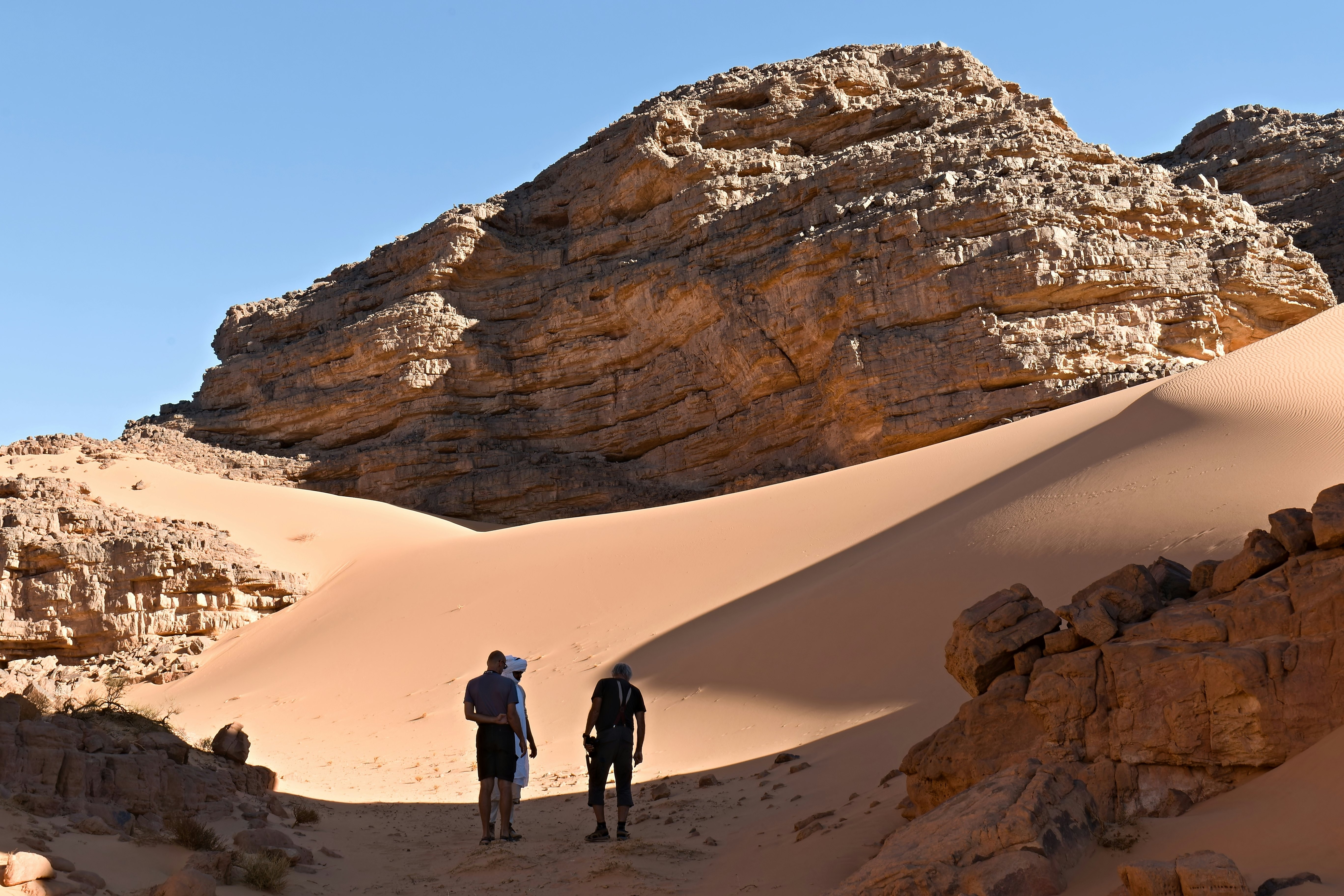 Two men in short pants walk on reddish sand behind a guide wearing all white and a head wrapping toward a rock formation in Algeria.