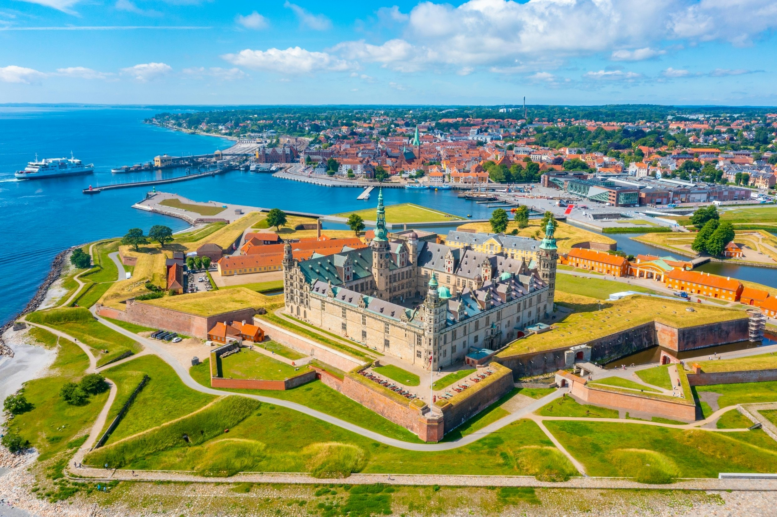 Panorama of a large castle surrounded by green grass, with a waterway and skyline of low, red-roofed buildings in the distance on a bright, sunny day.