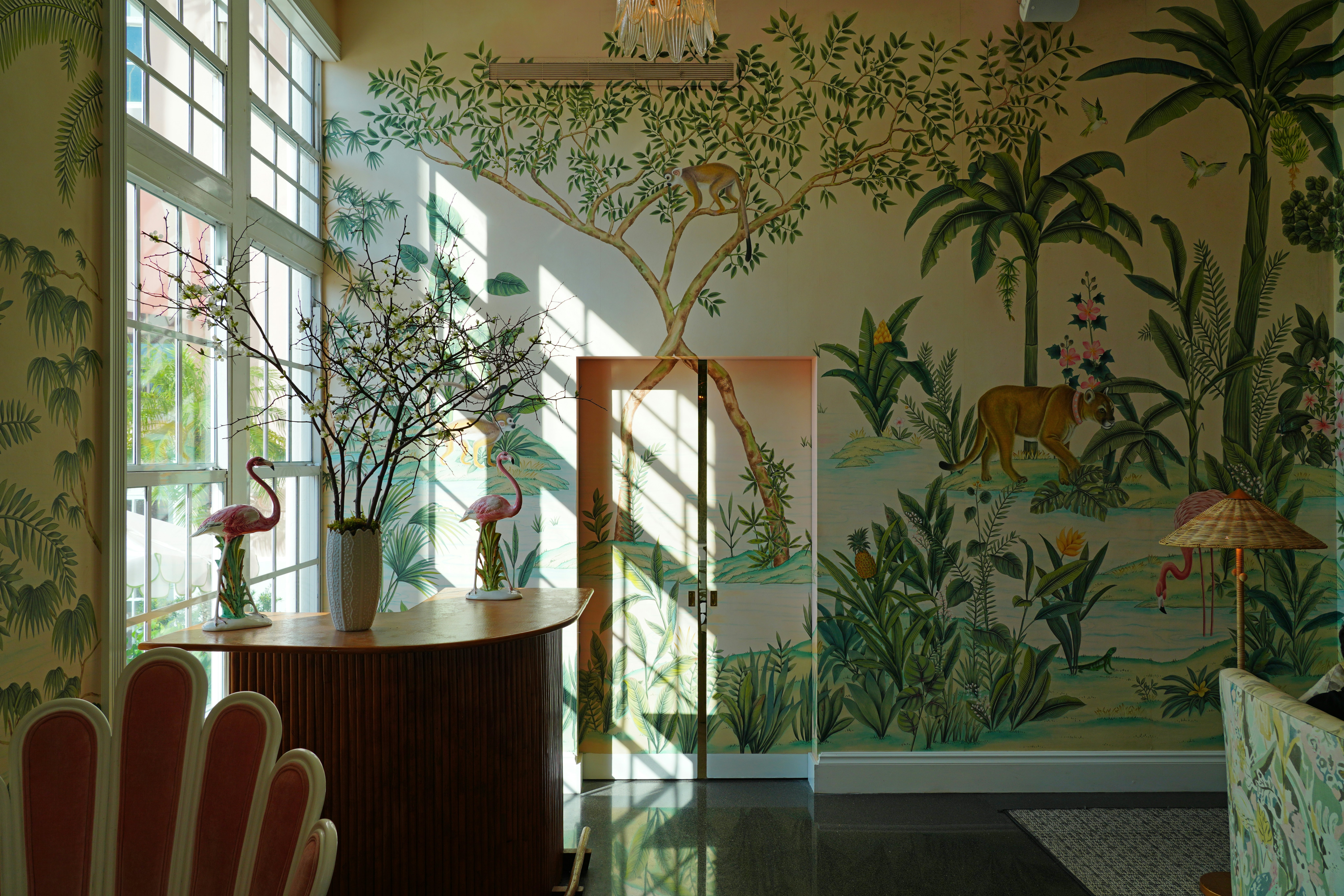 A room with a floor-to-ceiling window on one side has wallpaper with a green tropical print; there is a tall dark wood desk with sculptures of pink flamingos.