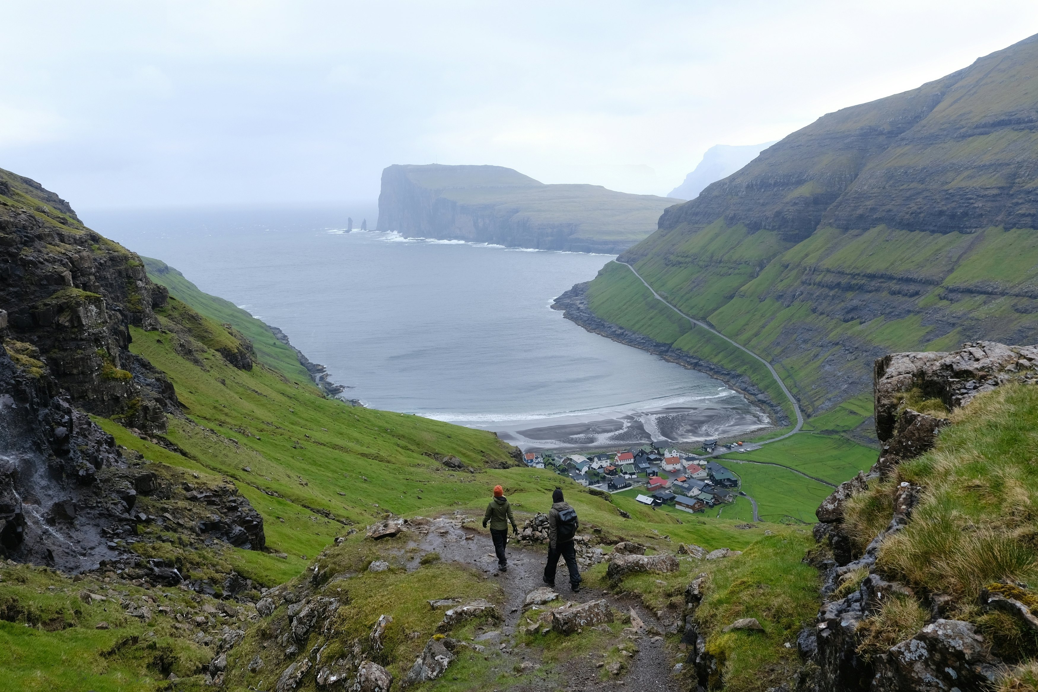 Two hikers in wet-weather gear follow a coastal path down towards a small settlement at the foot of green cliffs.