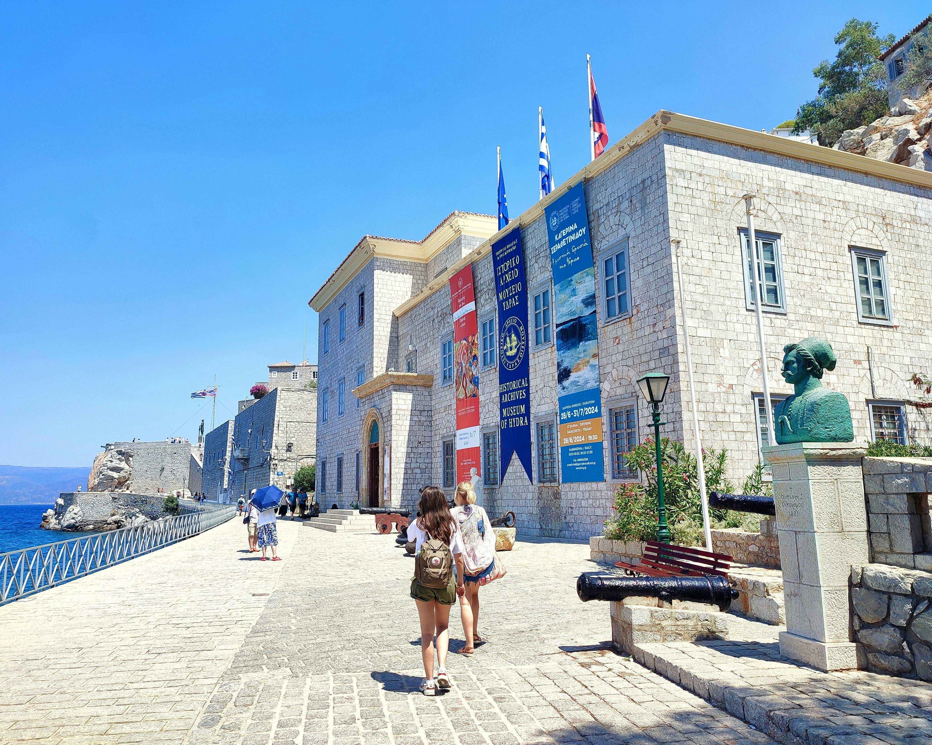 People walk on a cobblestone promenade by a harbor past a bronze bust of a man.