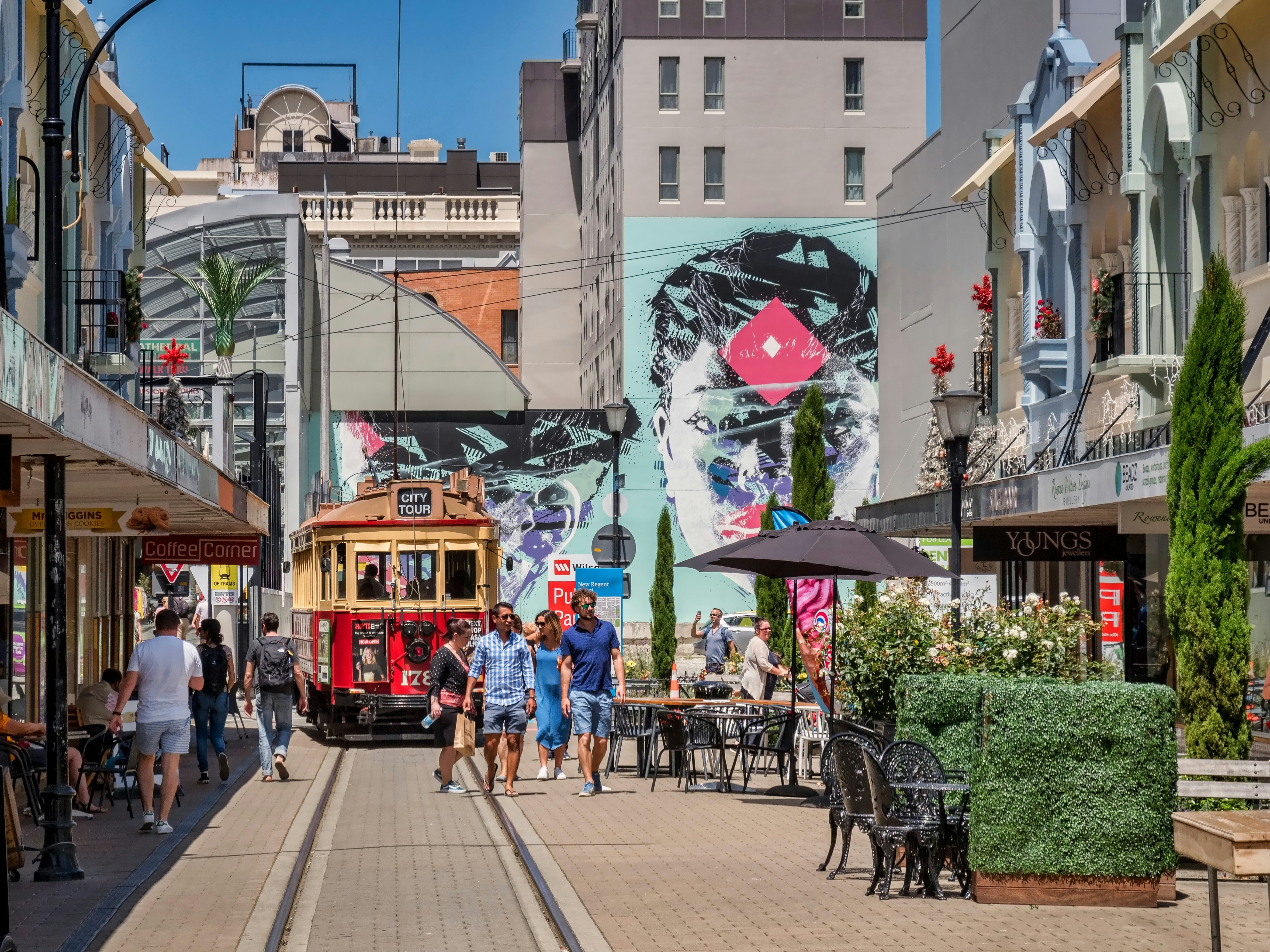 People walk past outdoor cafes and shops on a street in New Zealand with a tram route running through it.