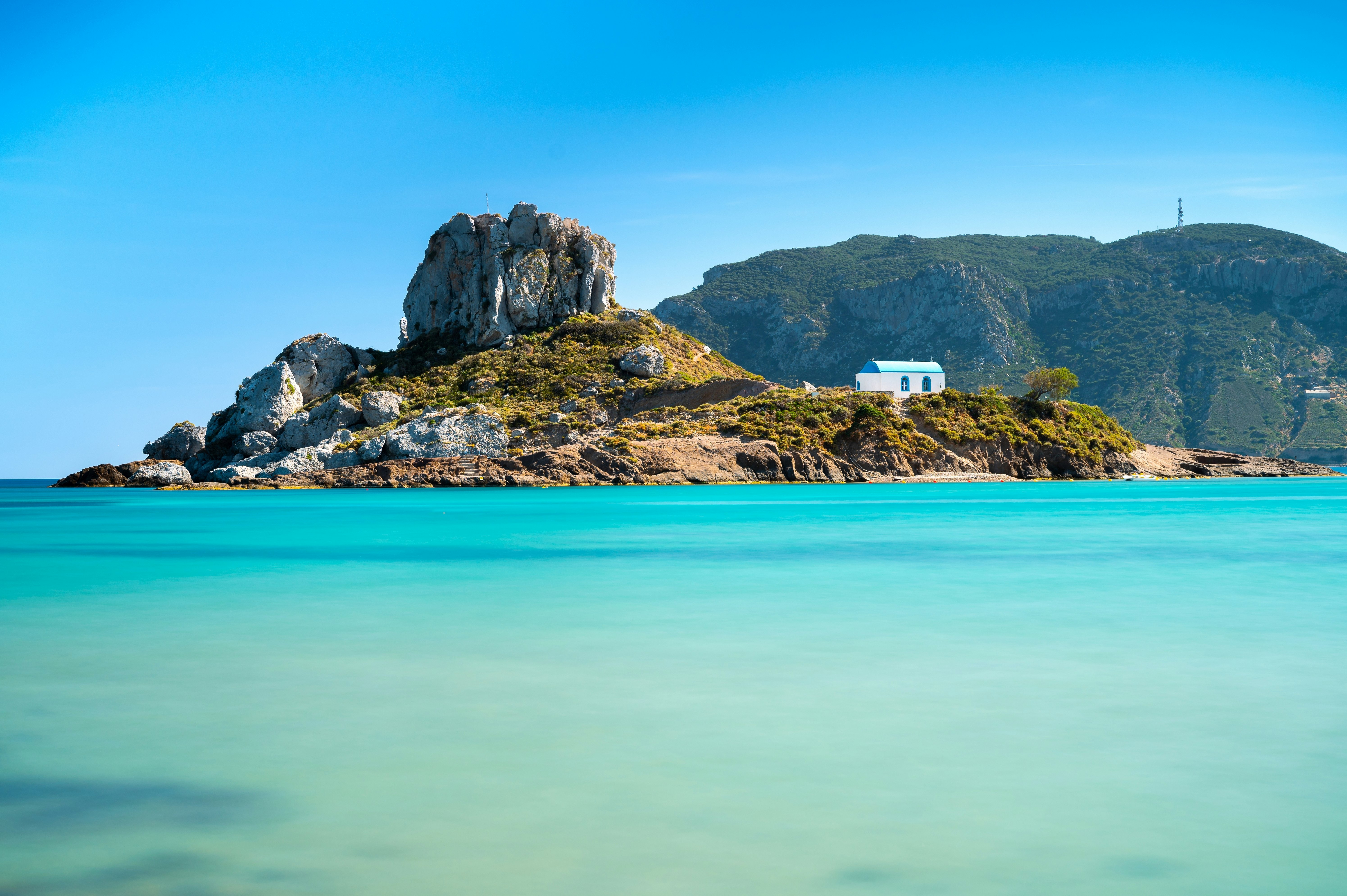 A rocky islet with a small white church is pictured against an aquamarine sea.