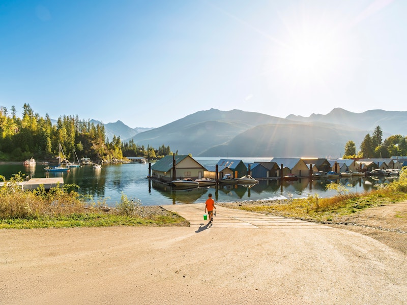 A young boy with a fishing rod and tackle box walks on a dirt road toward a bay boat ramp and boathouses along a lake on a sunny day.
