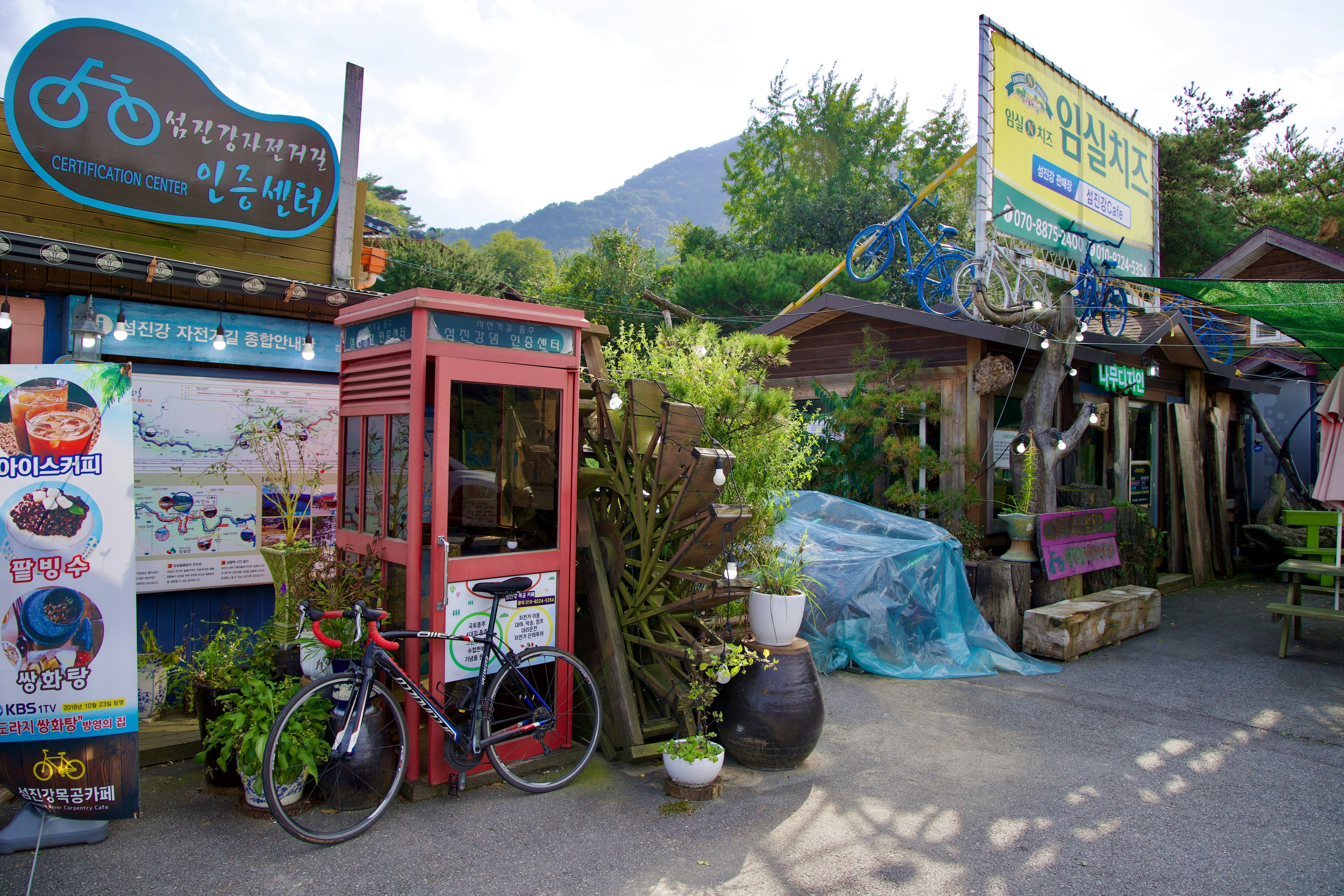A red booth with a bike leaning against it next to a water wheel various plants and a little shop topped with decorative bicycles.