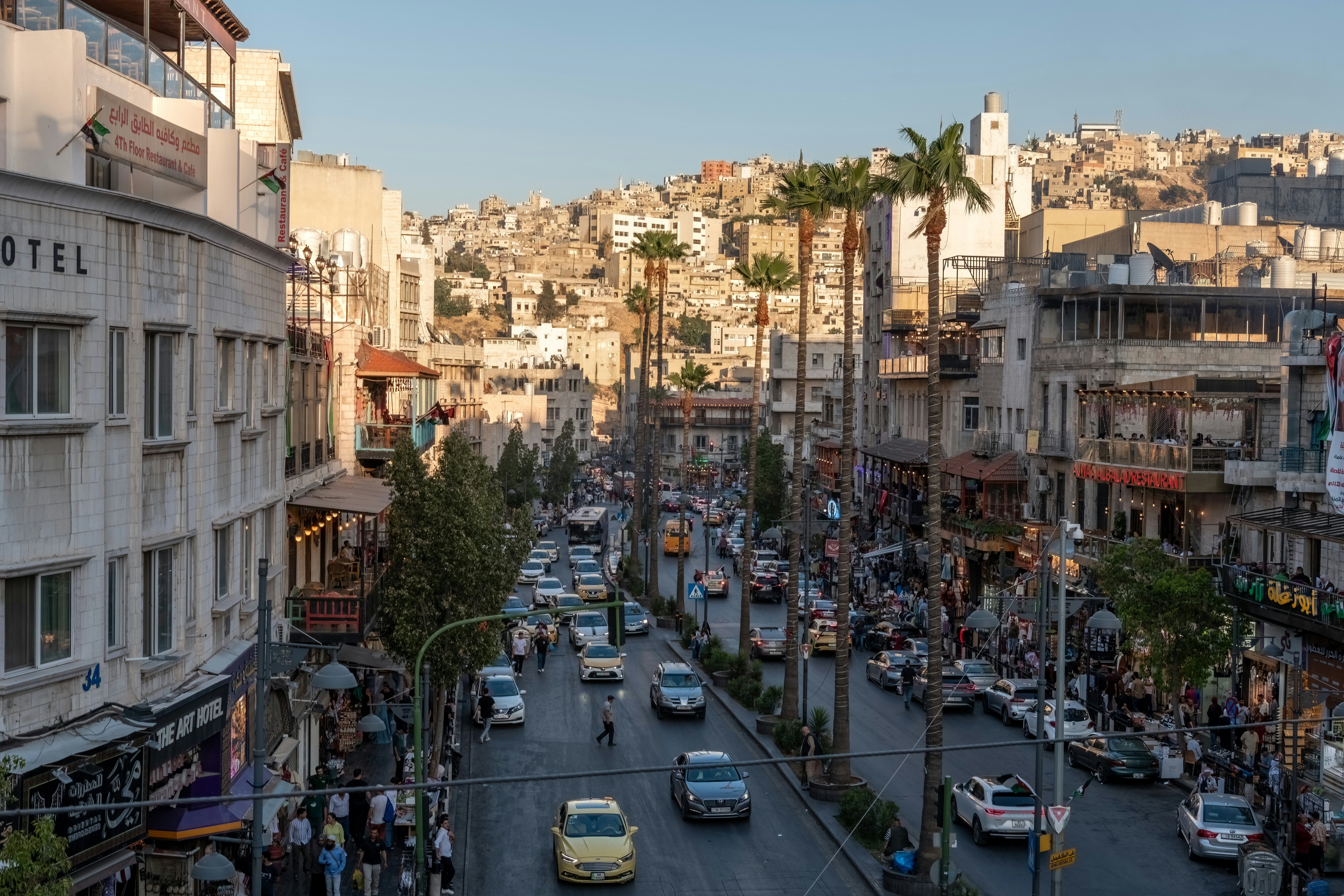A busy city neighborhood with people and cars. Cafes and shops line the street.