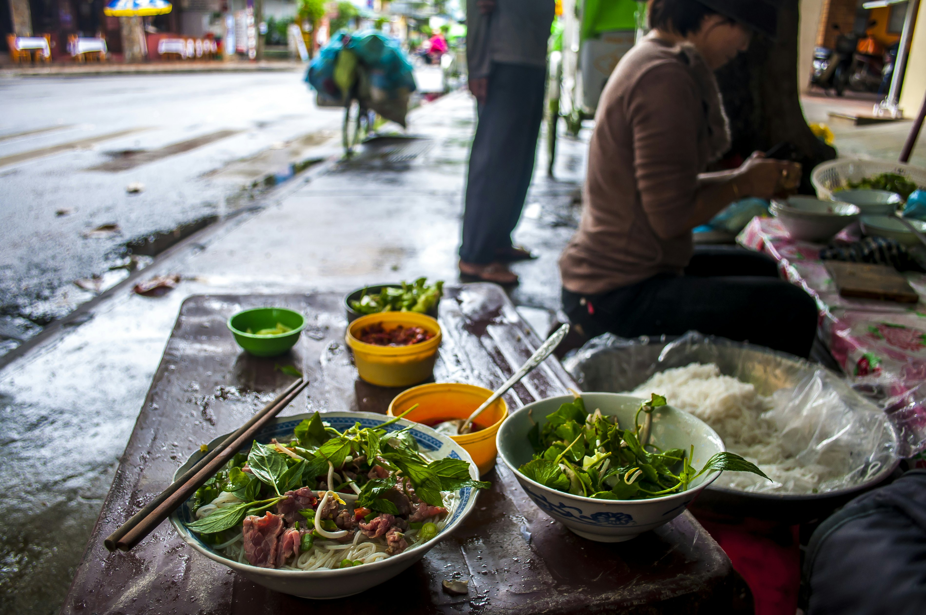 A food stall beside a street on a damp day. Bowls of noodles and vegetables sit on the table.