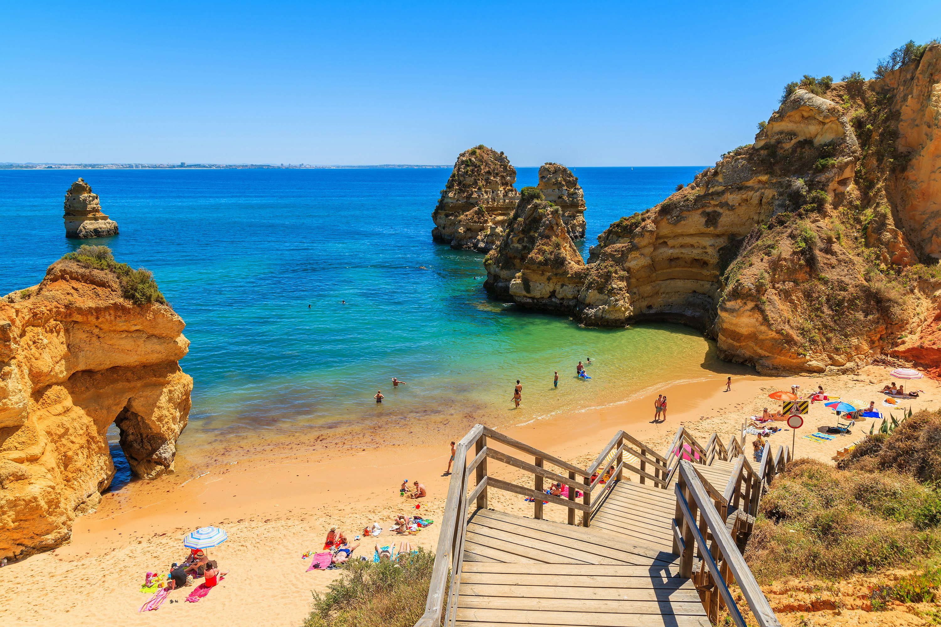 Wooden steps lead to a cove in Lagos, Portugal, where people are on the sand and in the water; steep rock formations rise on either side of the beach.
