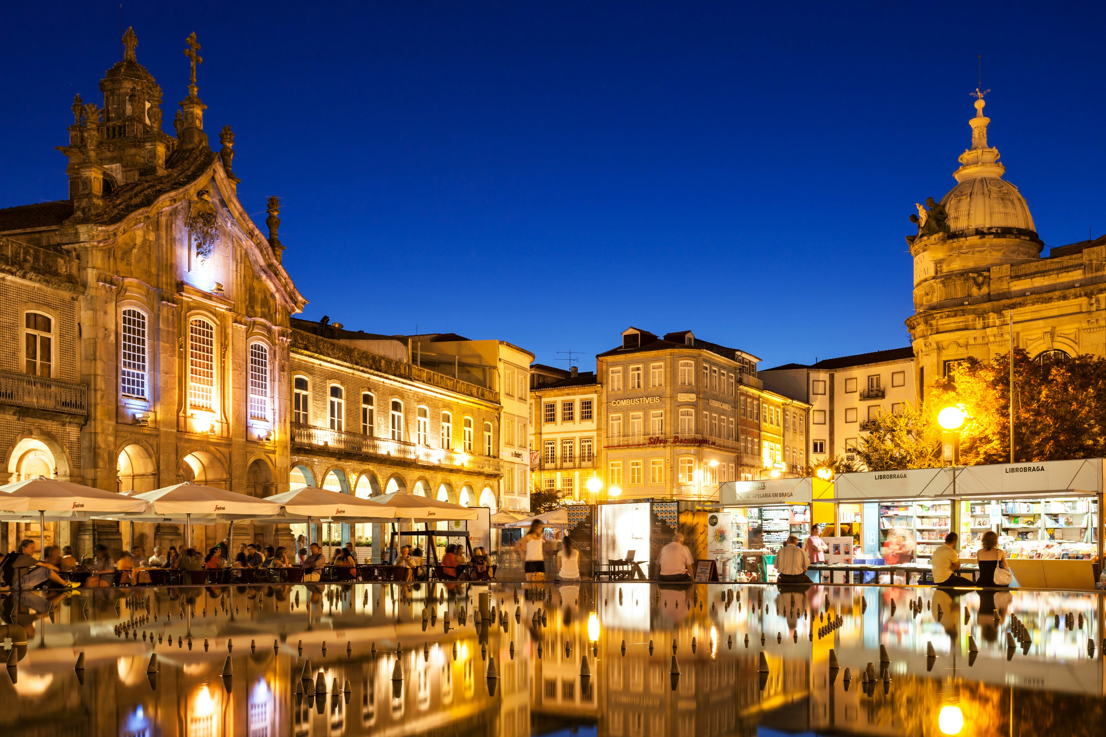 A city square in Portugal, pictured at night, is surrounded by stone buildings and has a water feature in the center; people sit at a cafe under umbrellas at the edge of the basin, and there are stands on the other side.