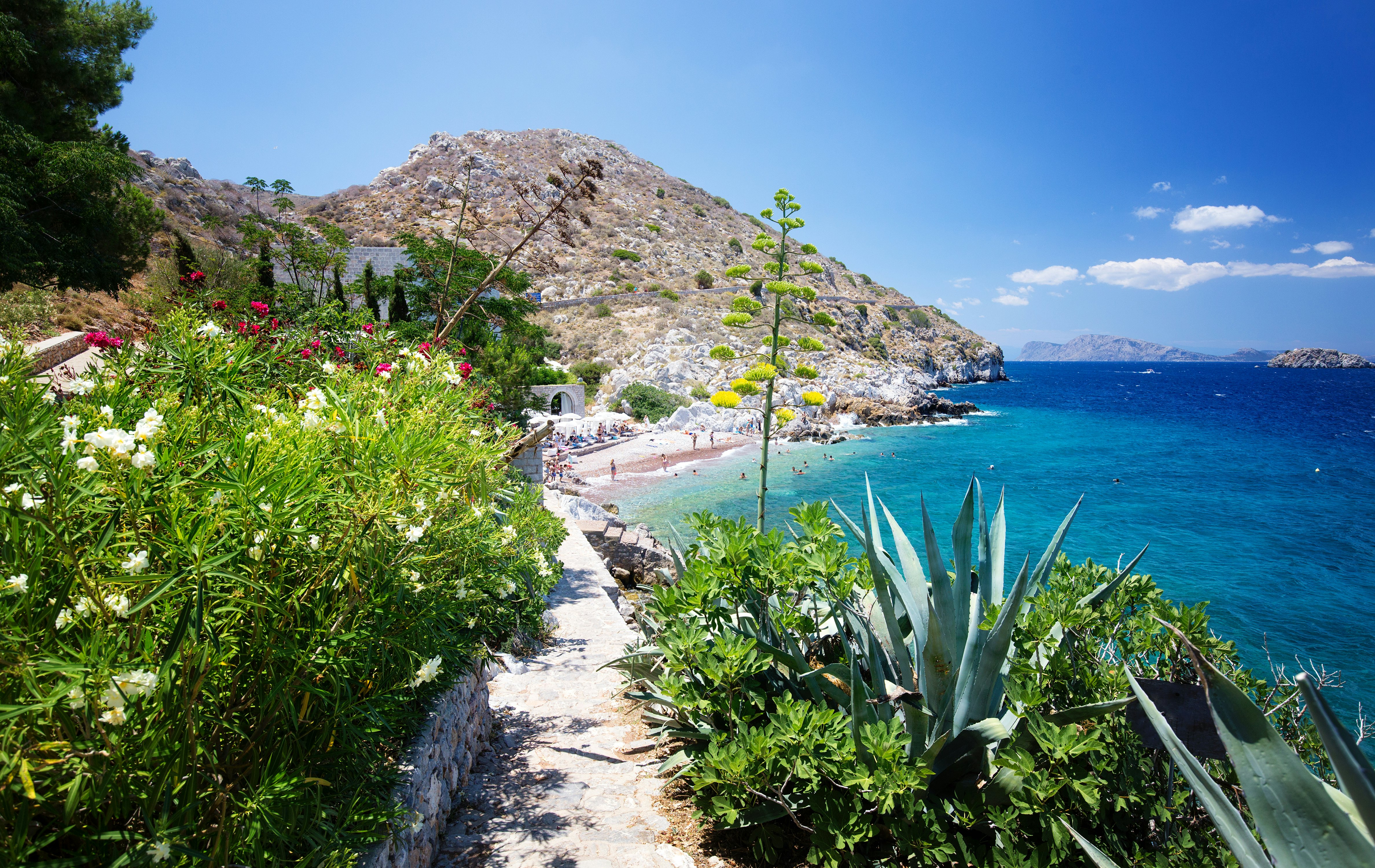A waterfront path leads past bushes toward a rocky beach.