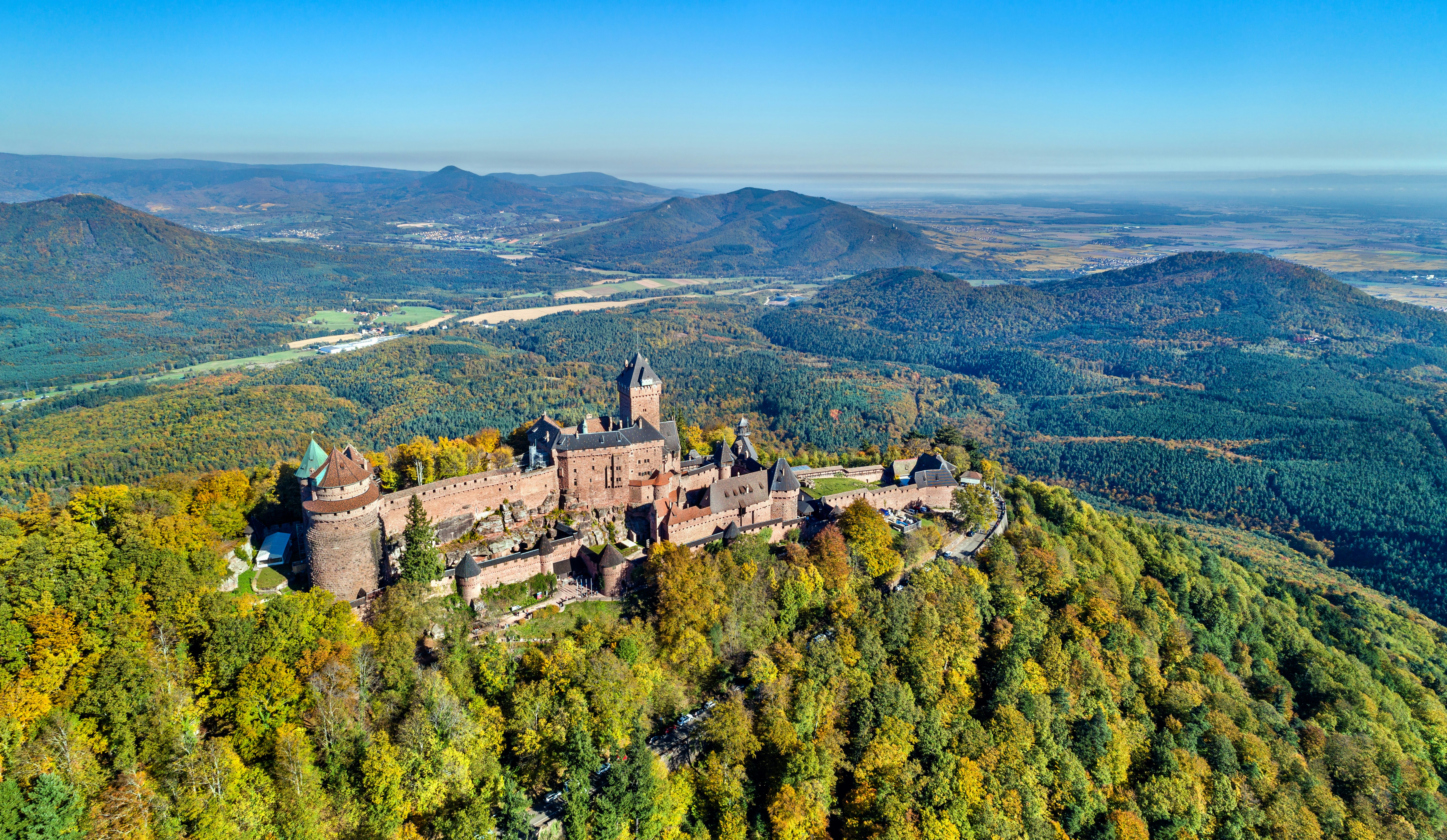 A hilltop chateau surrounded by woodland that is changing from green to yellow as the seasons shifts to autumn.