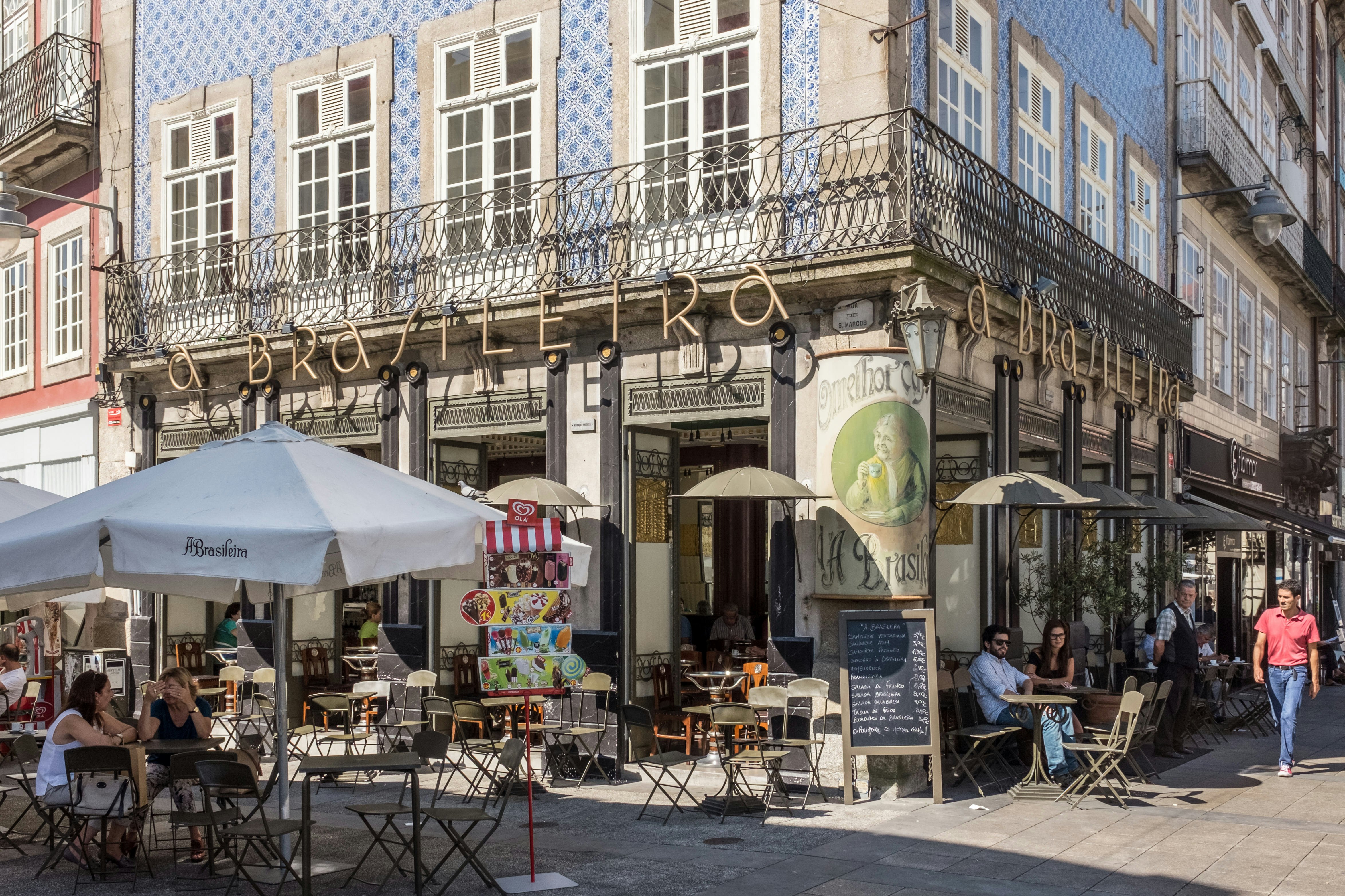Outdoor tables are set up on a sidewalk in front of a cafe at the base of building covered in painted blue tiles.
