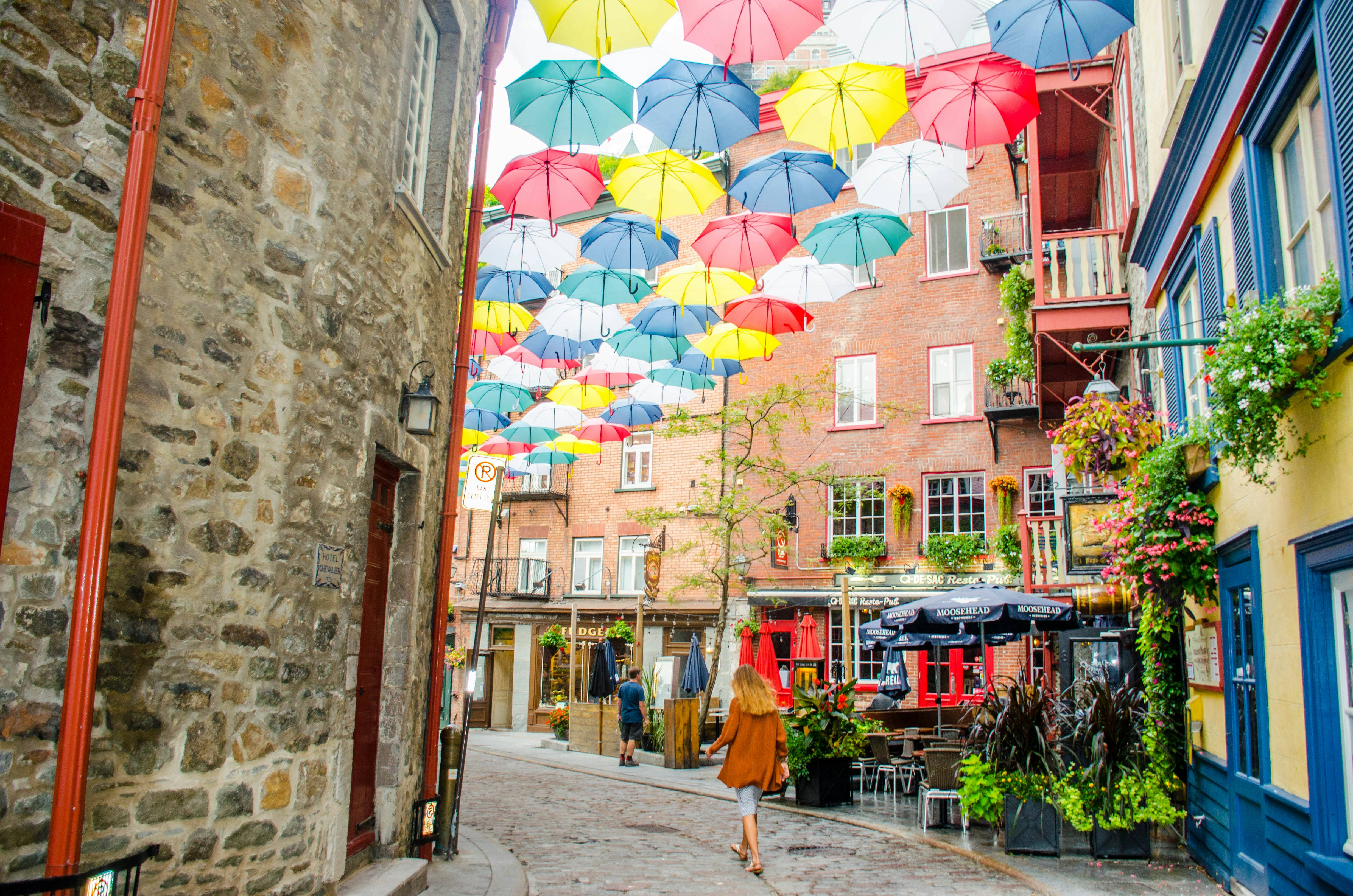 A woman walked on narrow cobblestone street surrounded by stone and brick buildings with colorful umbrellas suspended above on an overcast day.