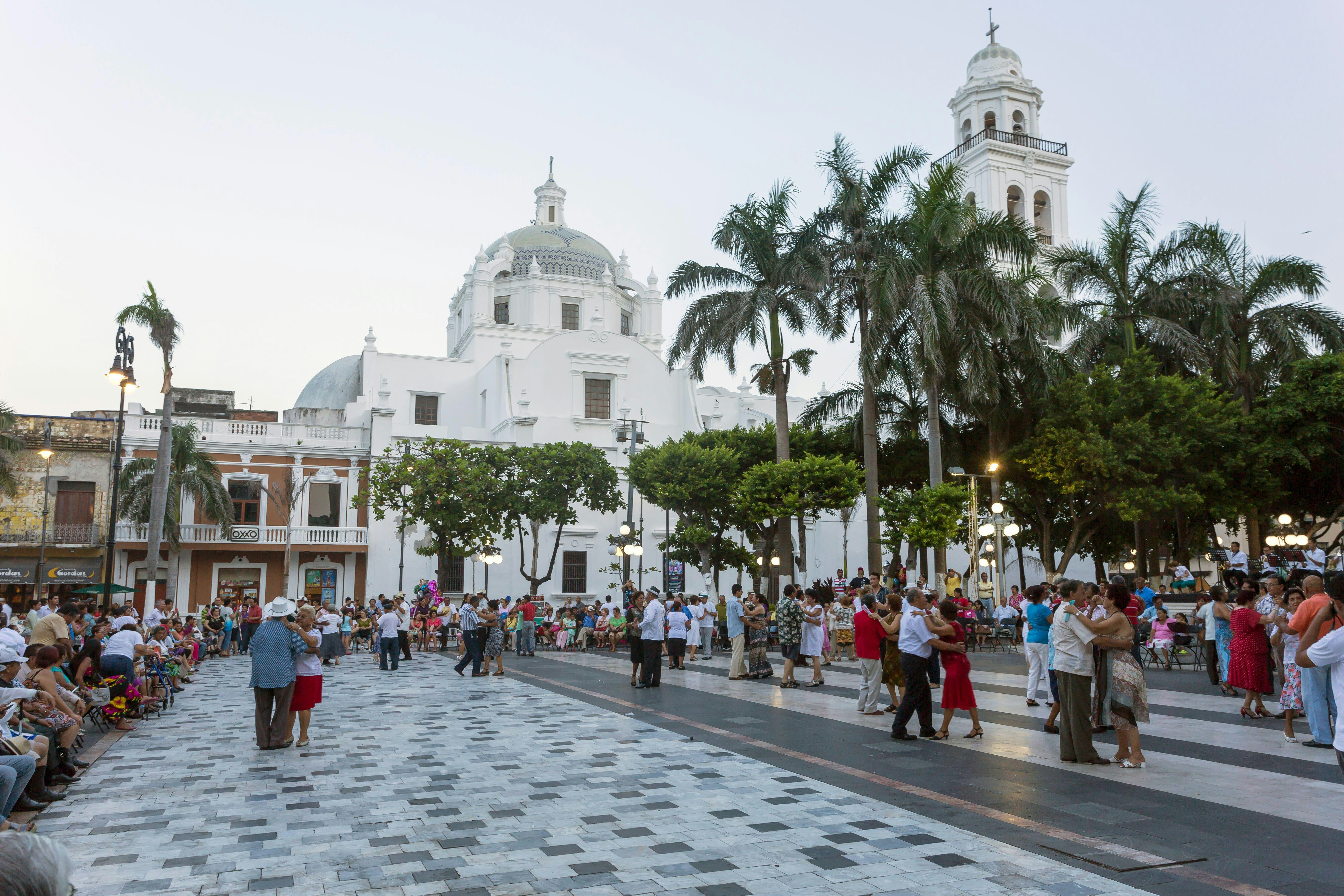 In the evening, couples dance in a city square with a large white building with a dome in the background.