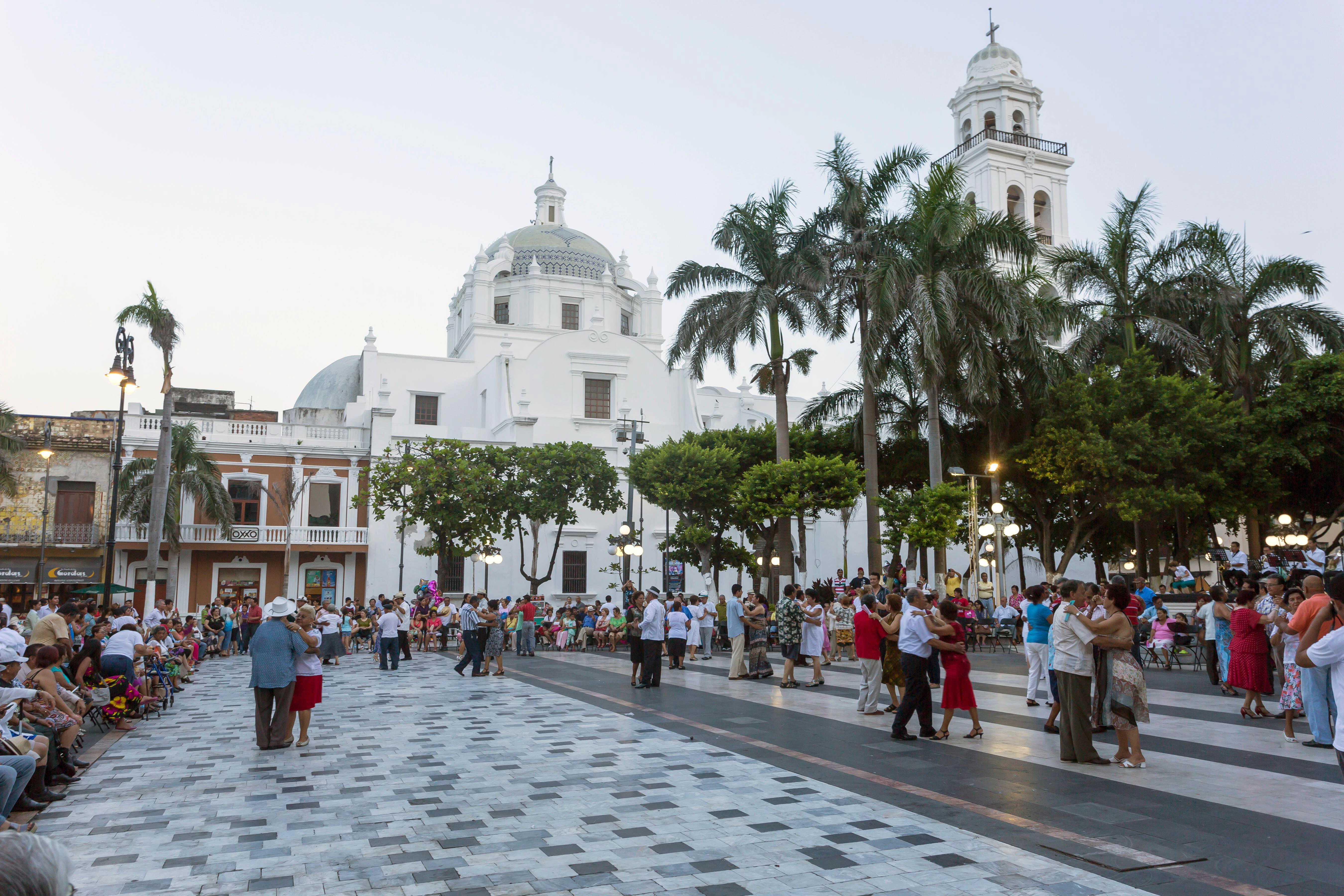 In the evening, couples dance in a city square with a large white building with a dome in the background.