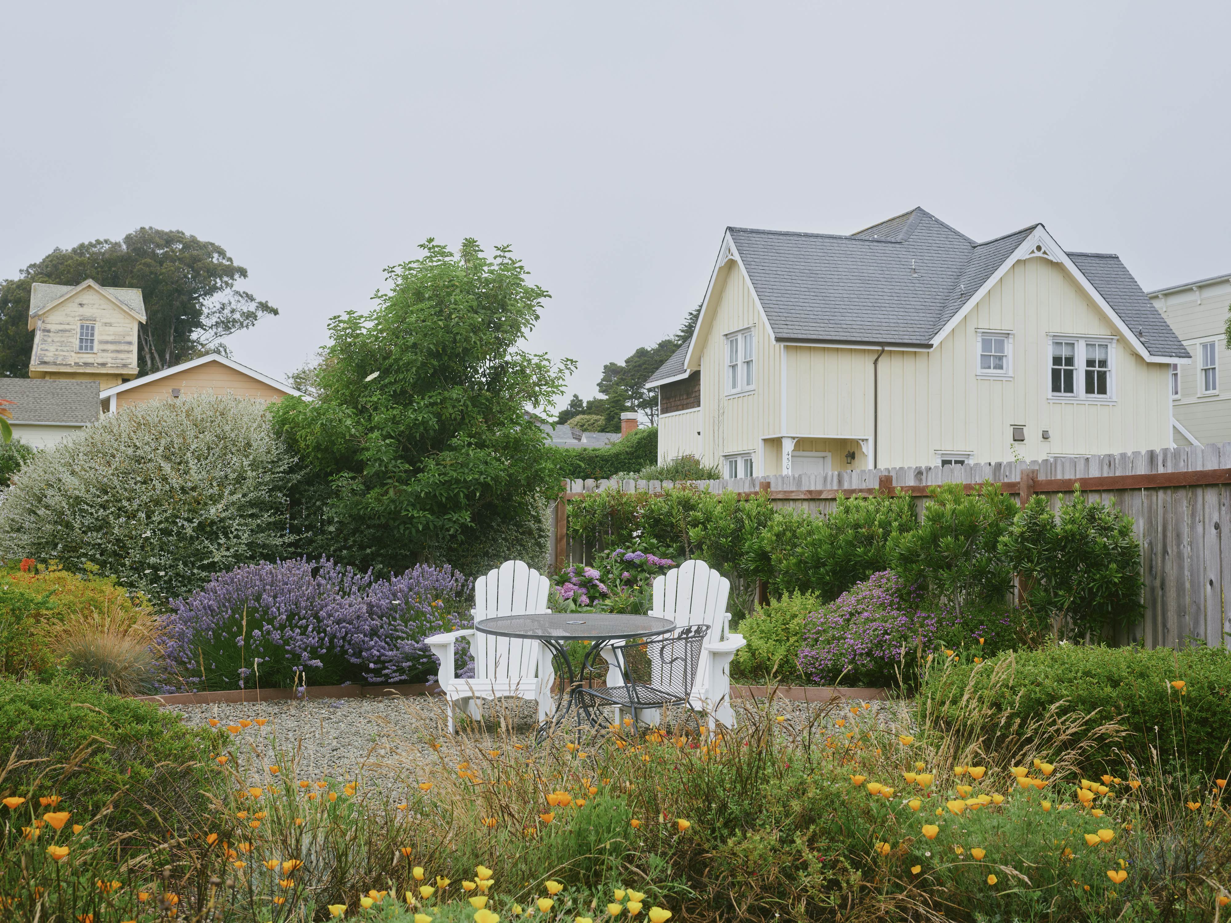 Two Adirondak chairs surrounded by flowering bushes in front of a yellow wooden house
