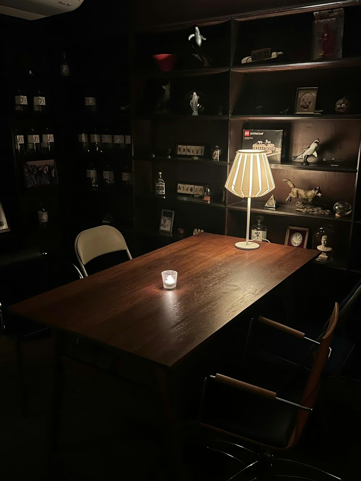 A dim room with dark furniture and a table lamp and a small candle lighting the room