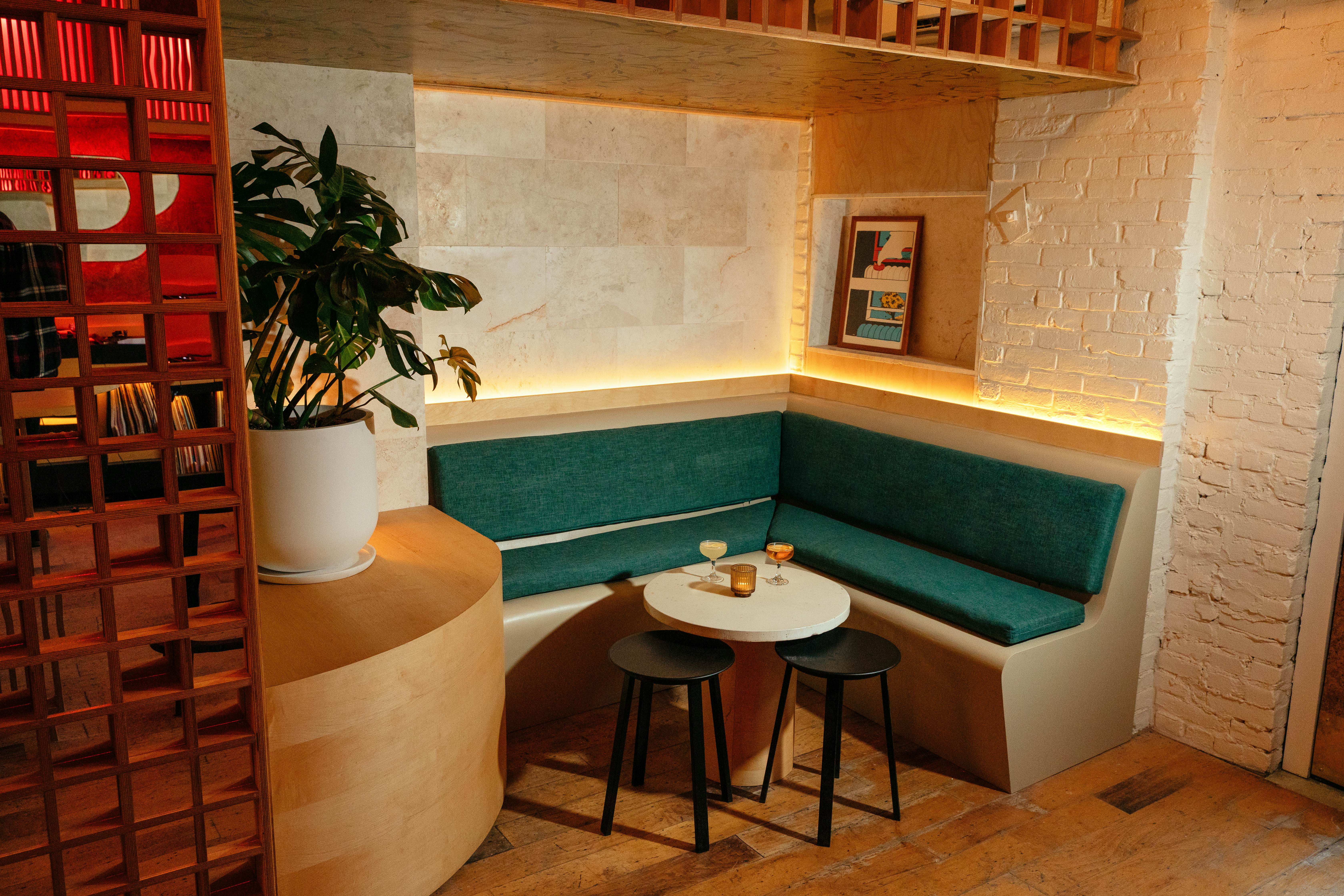 A beige corner booth with green padding faces a small round white table, under which are two black stools. There are three drinks on the table, and a plant on a shelf above the booth.