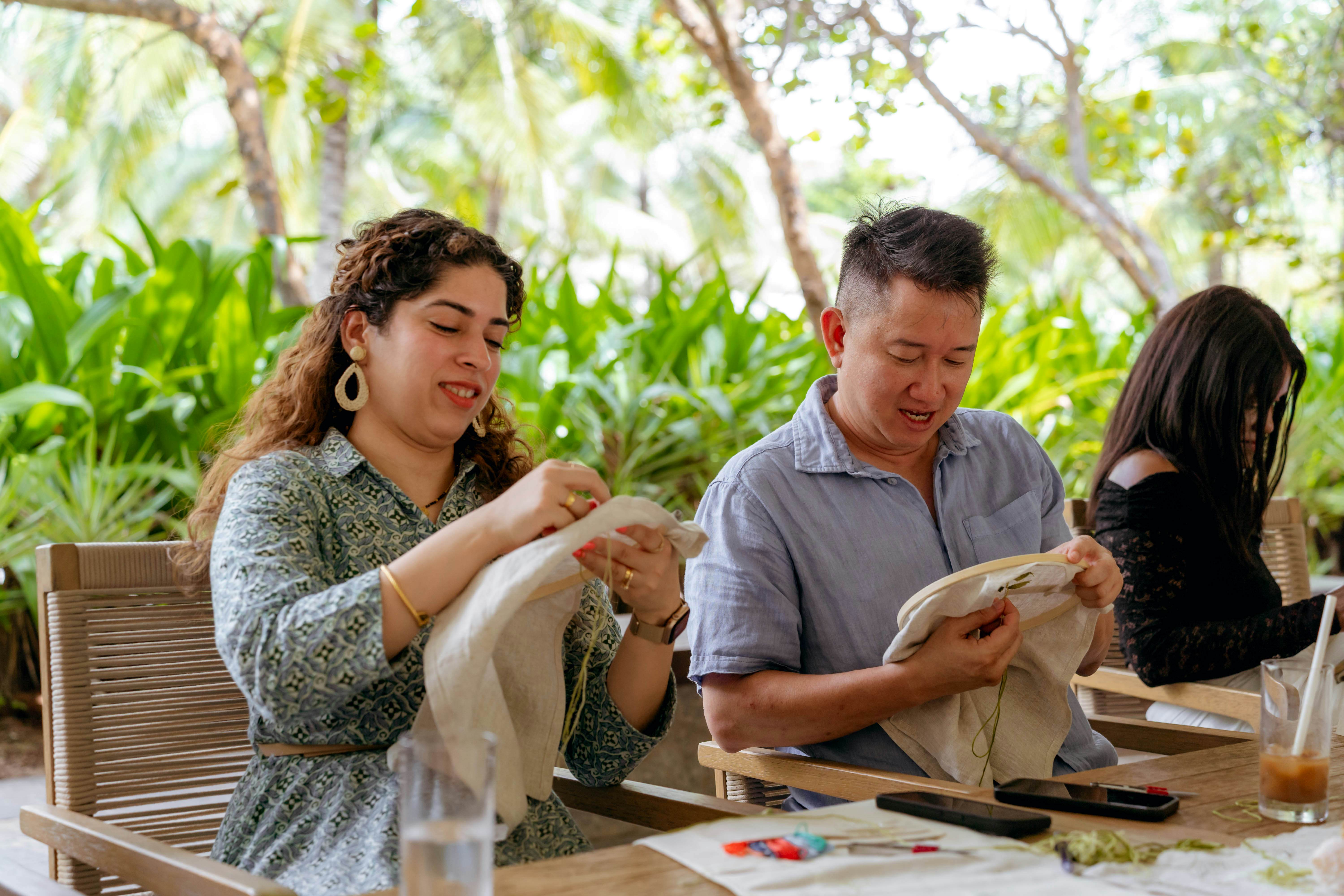 A man and woman sit in front of lush greenery with embroidery hoops in their hands.