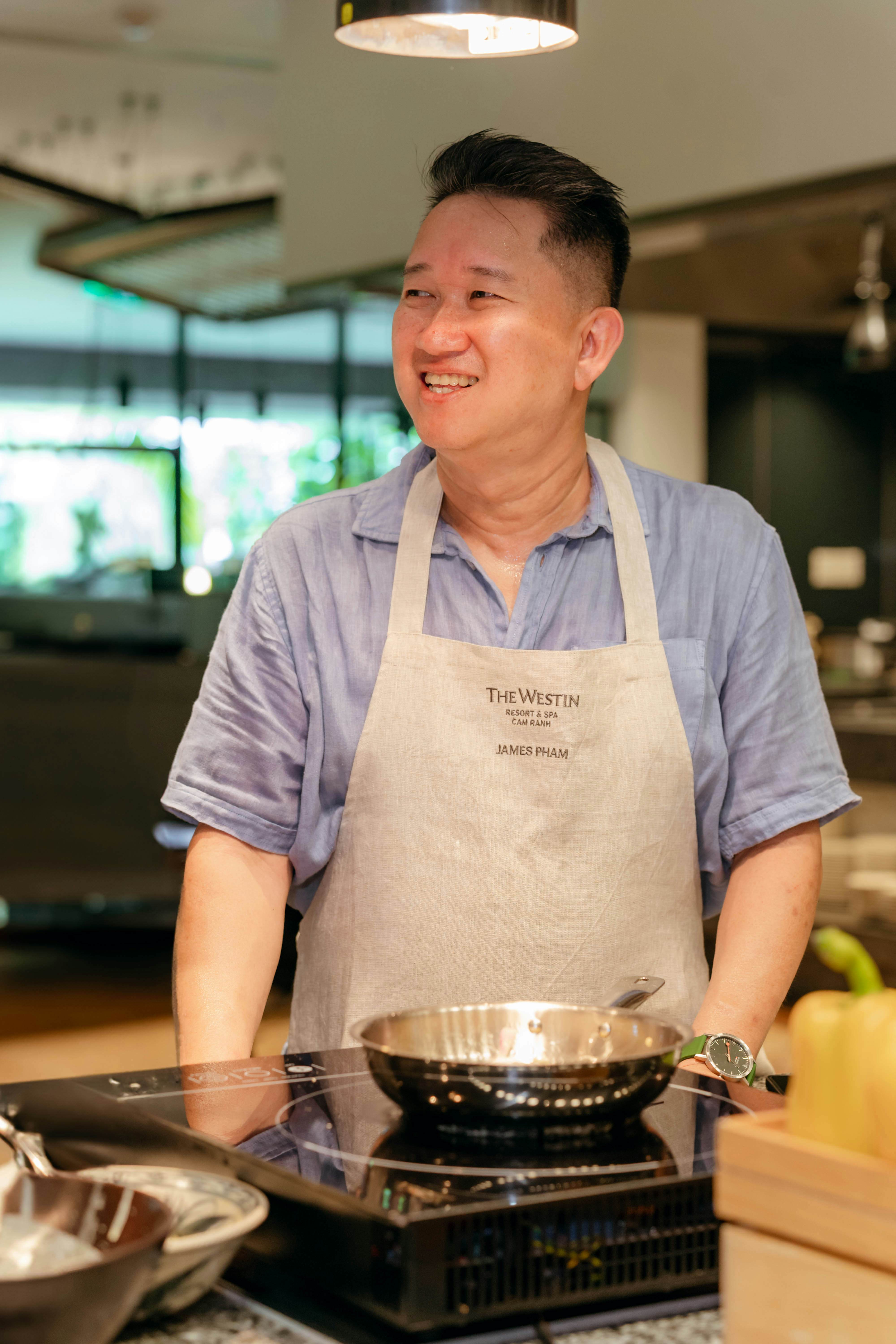 A smiling man wearing an apron stands in front of a countertop stove with a stainless pan.
