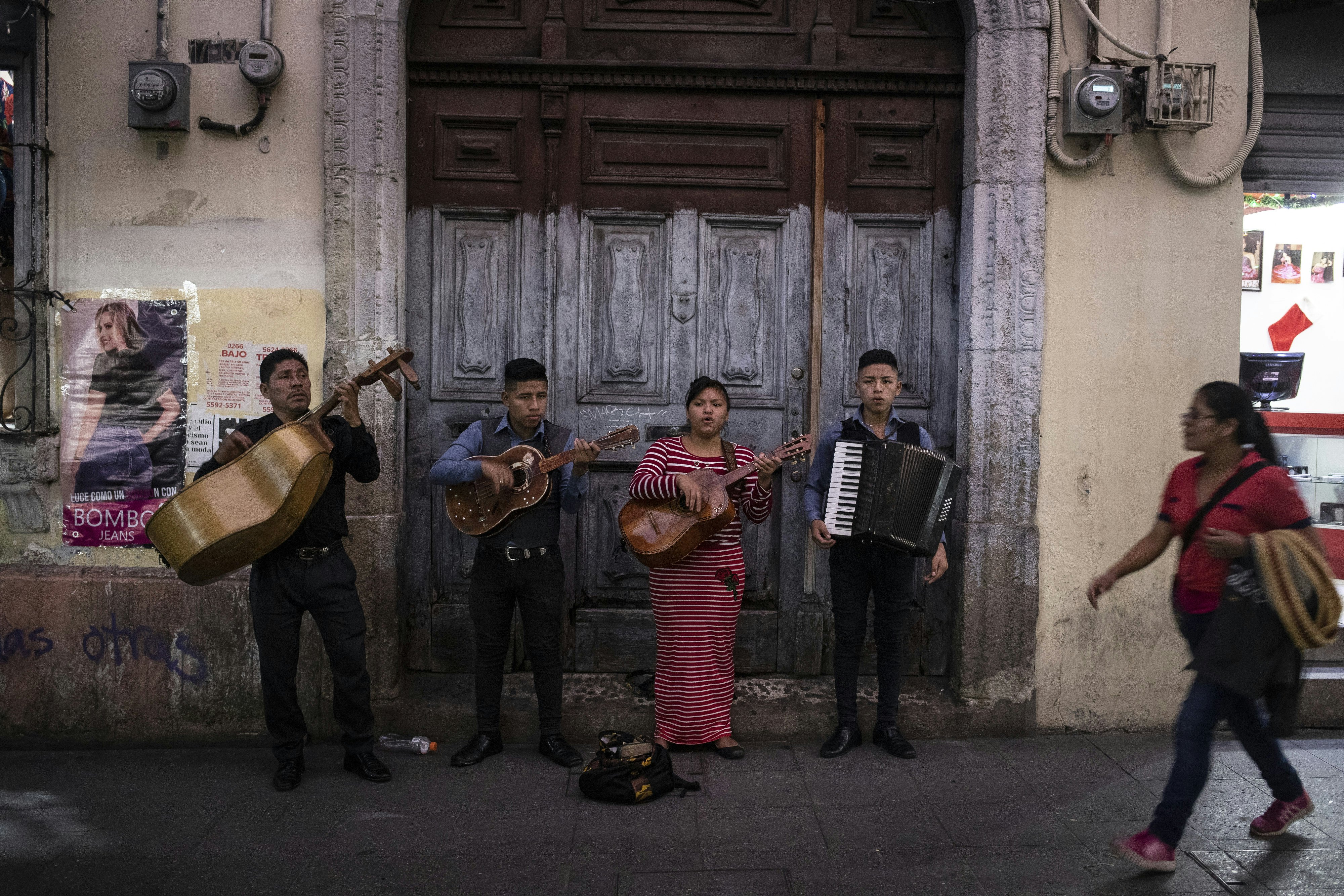 Mariachi musicians perform in front of a wooden door on a pedestrianized street in a city.