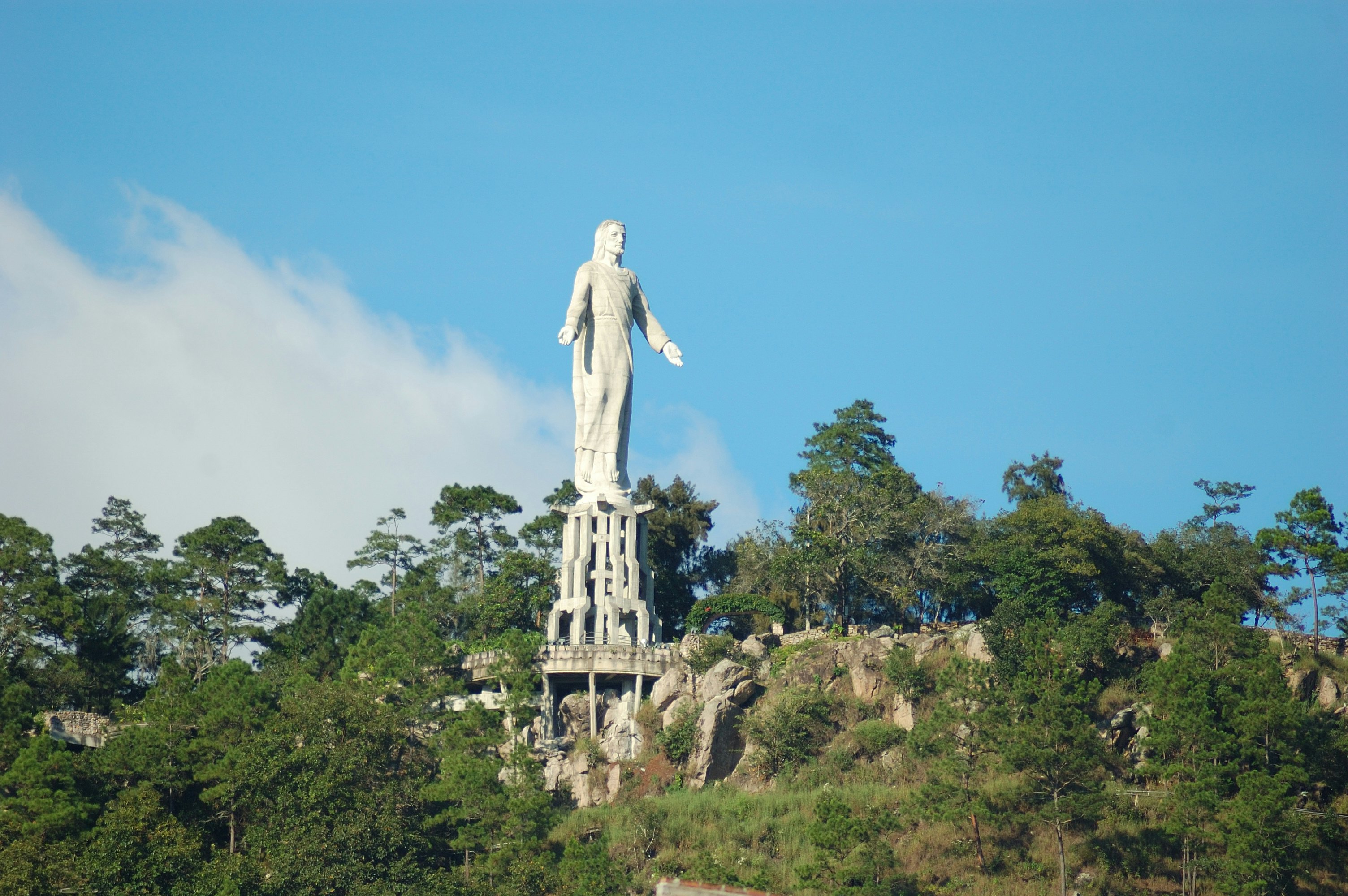 A stone statue of a man with his arms held open rises over the forests on a hilltop.