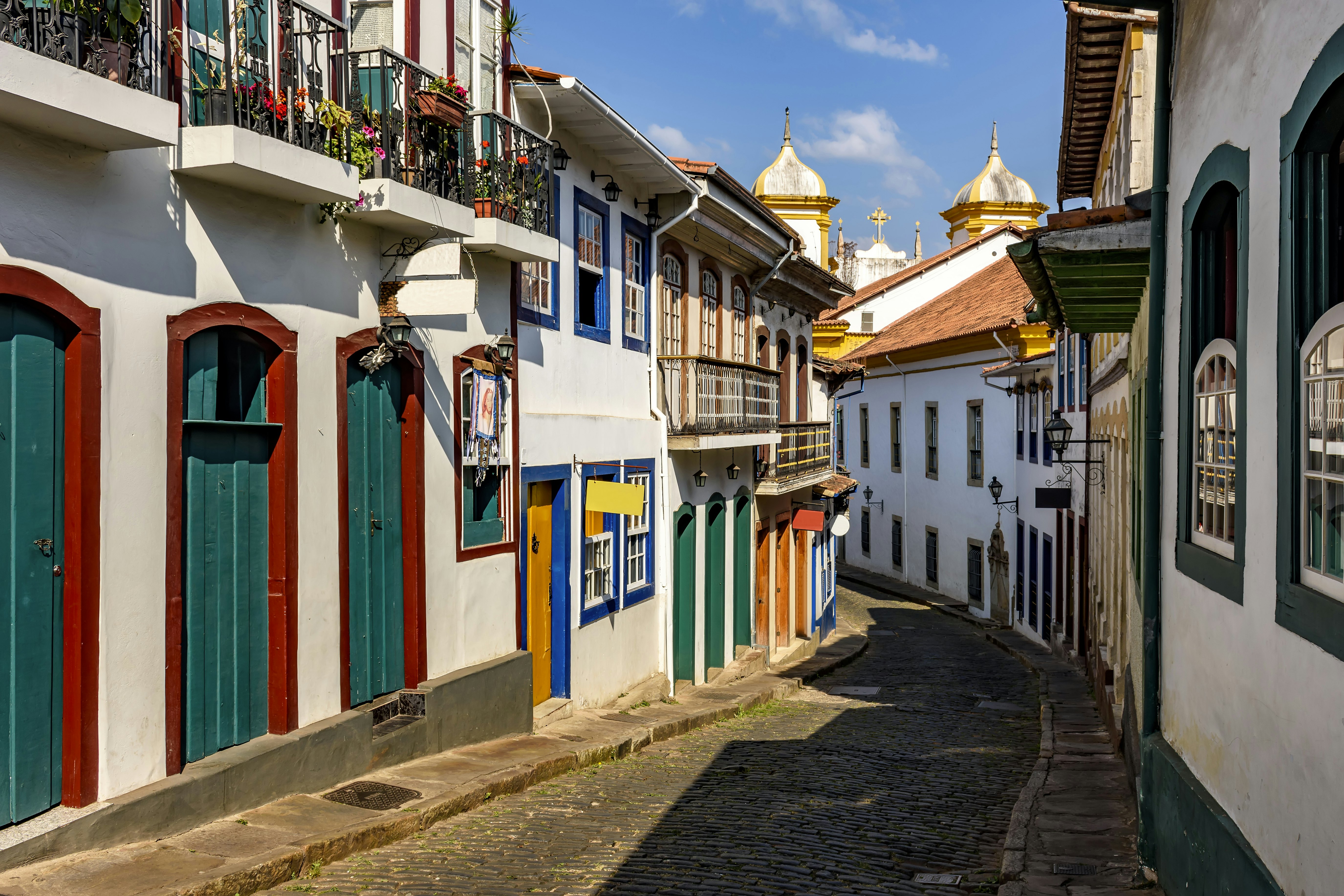 A narrow cobblestone street with white two-story buildings that have colored doors and shutters; the sun is shining on one side of the street, and the sky is blue with small white clouds.