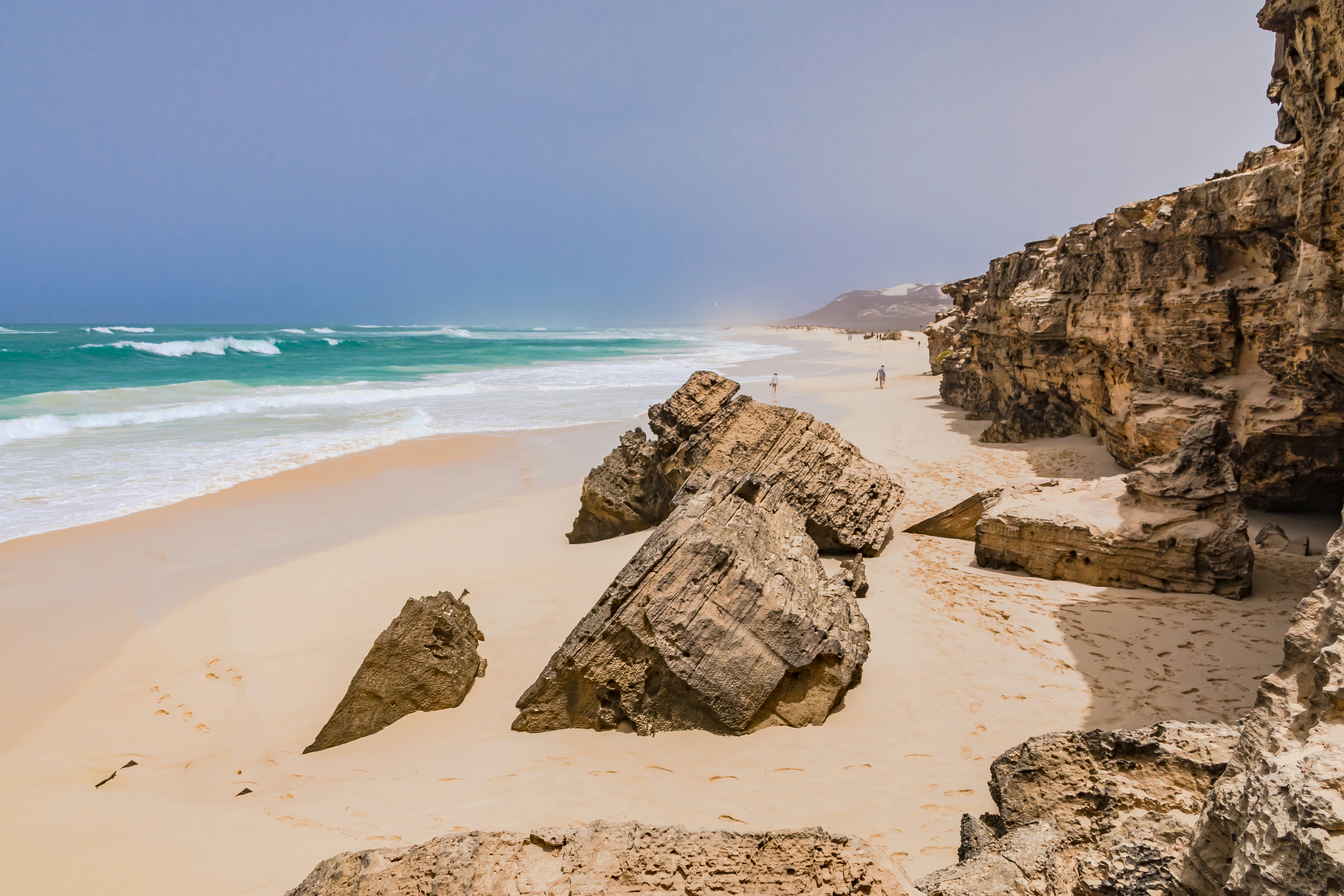 A view of a wide beach with rock formations. A wind picks up sand and creates whitecaps on the lapping waves.