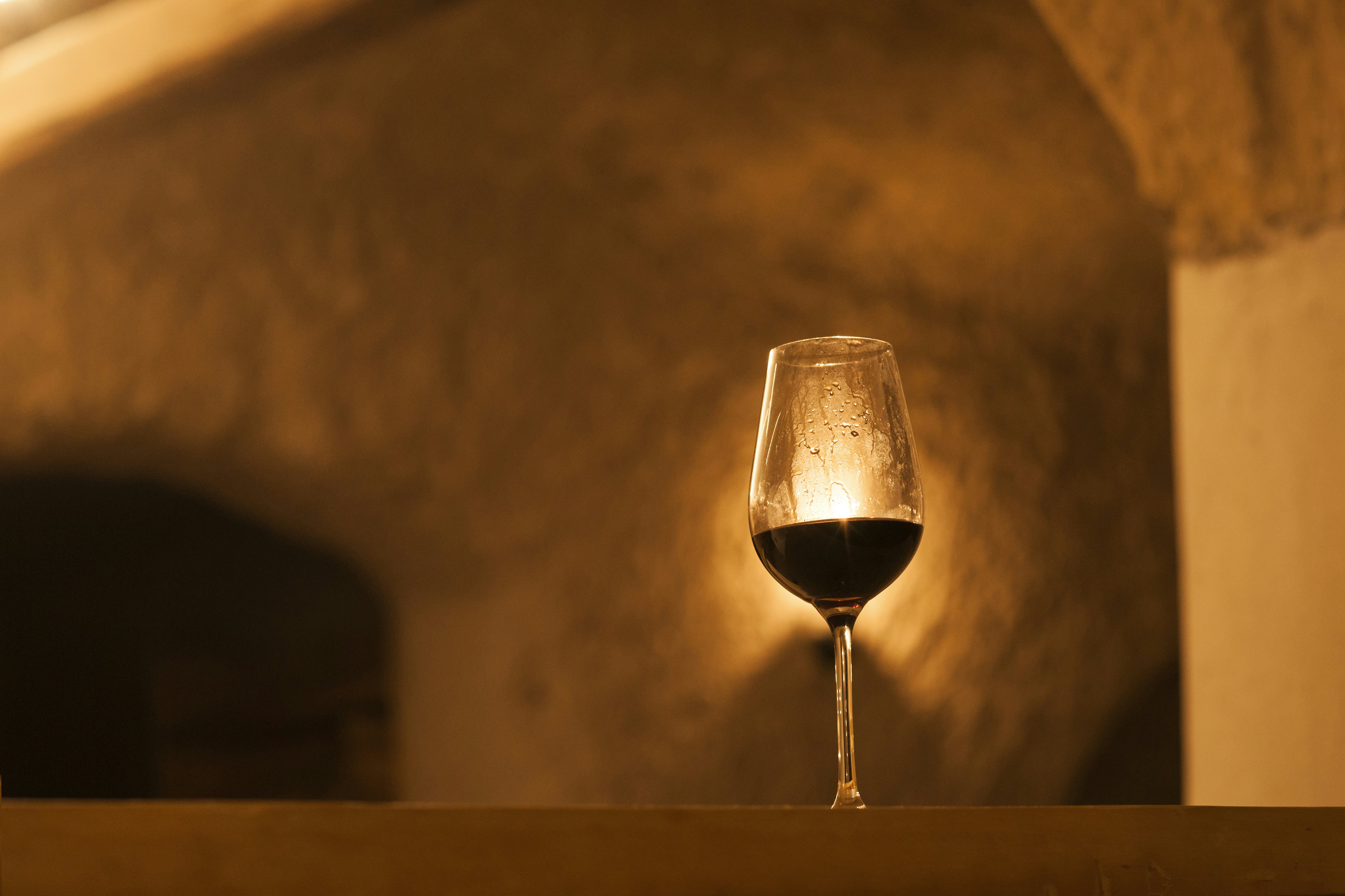 A glass of red wine on a shelf; the clear glass is lit from behind by a light on a wall.