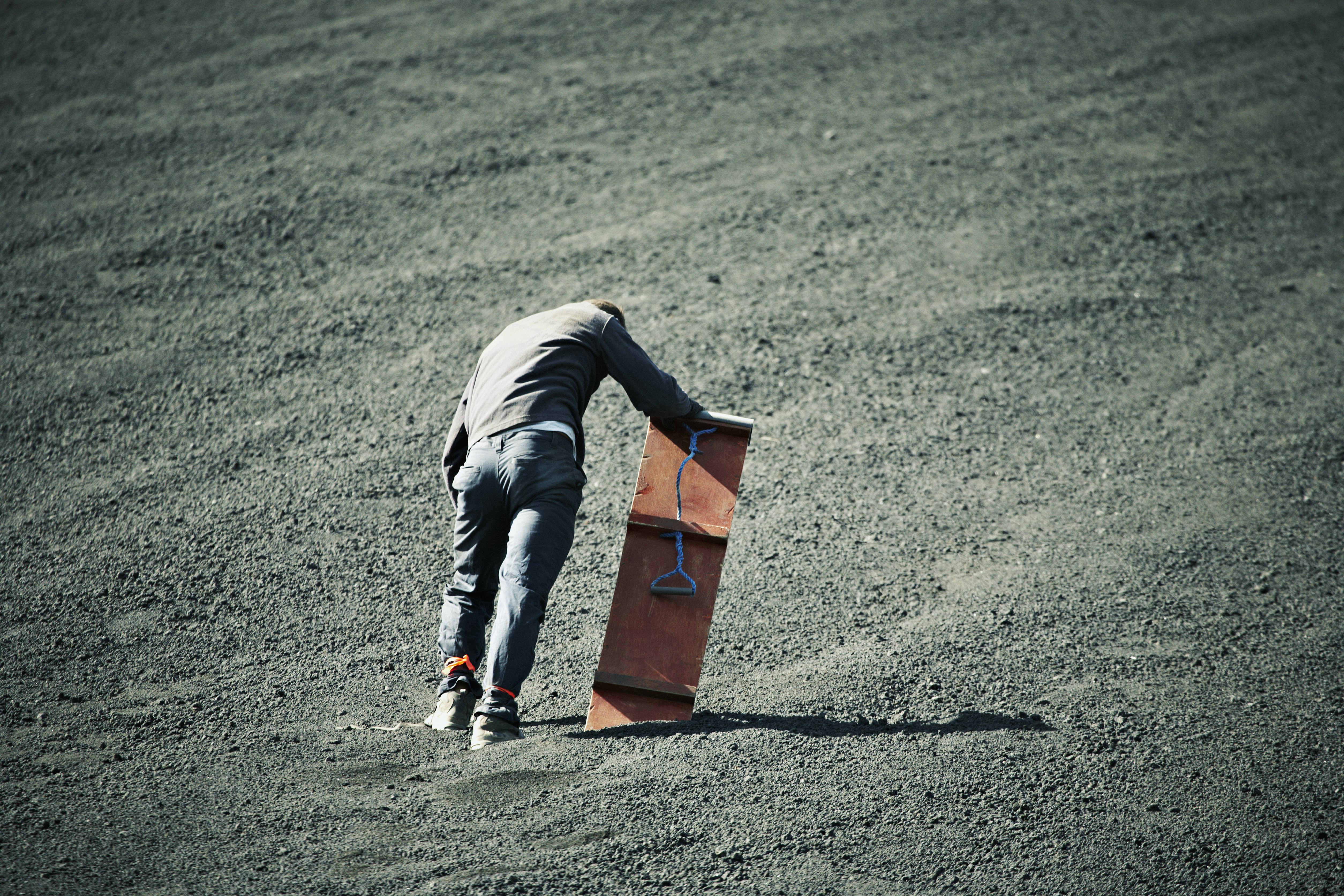 A tired man holding a sandboard on a dusty volcanic landscape.