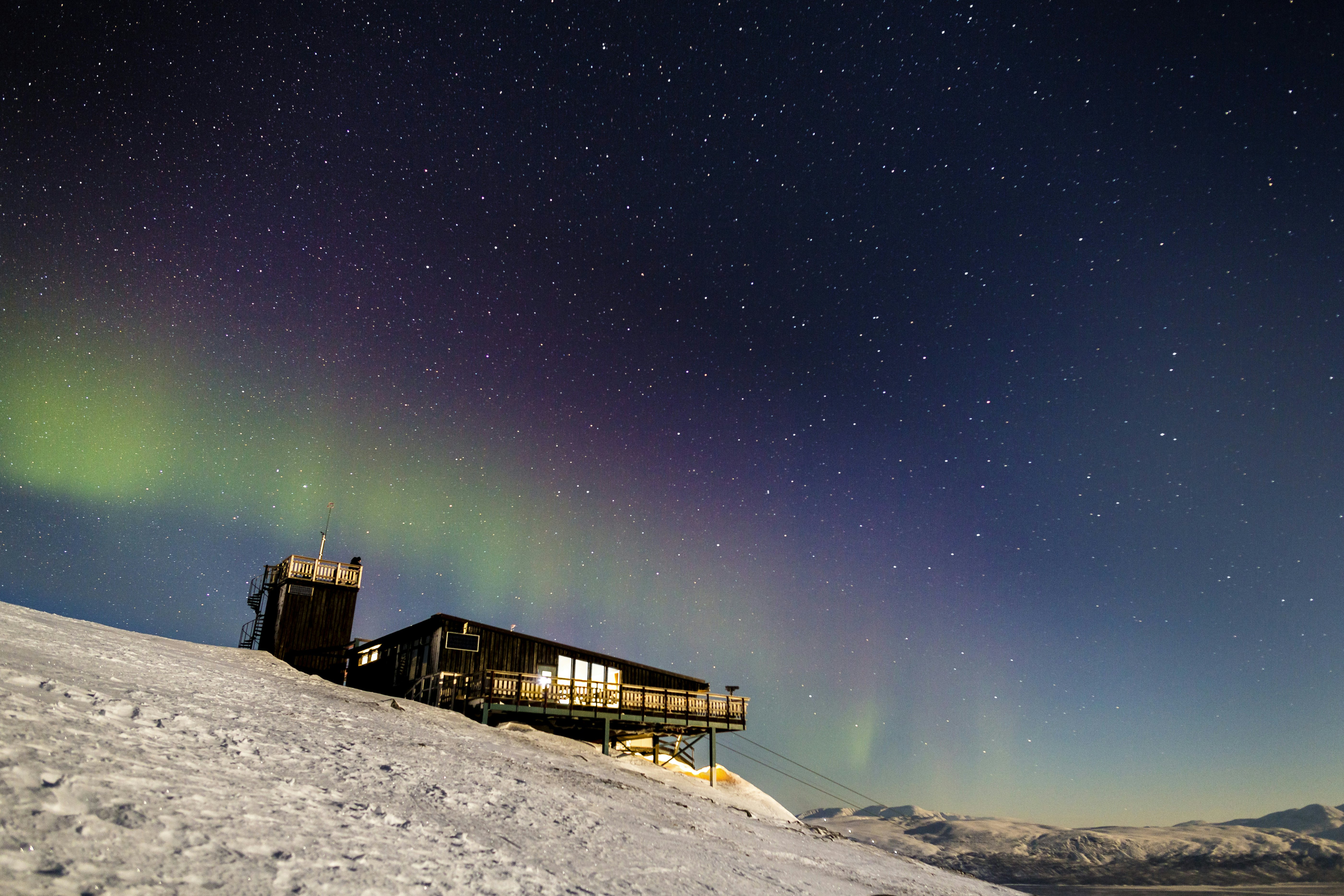 The northern lights and stars shine above a wooden building perched atop a snowy hill.