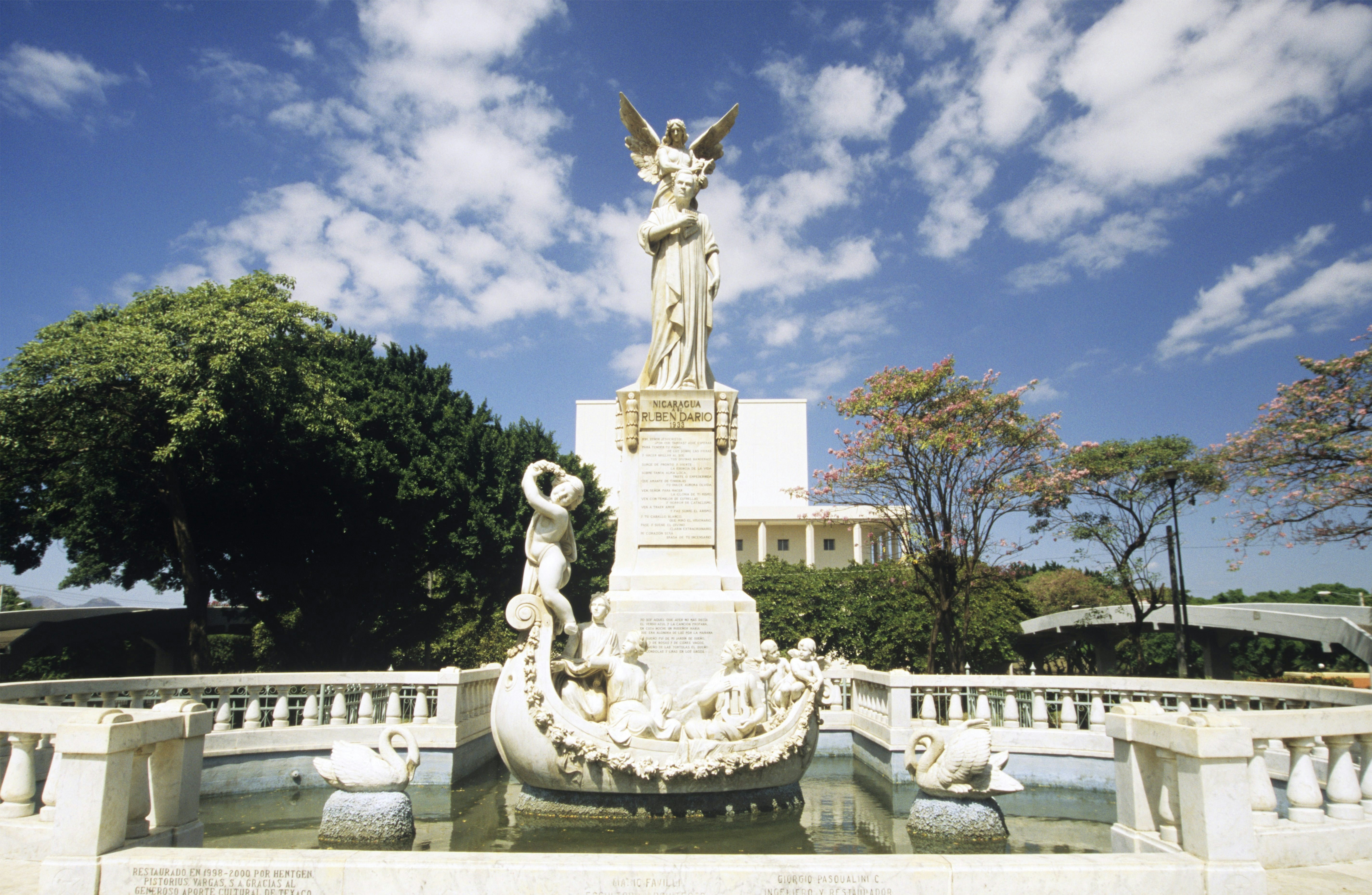 Fountain with a sculpture of a man in the middle. A sculpture of people inside a boat is in front of the man, and two swan sculptures are on both sides of them. A building is partially visible behind the fountain.