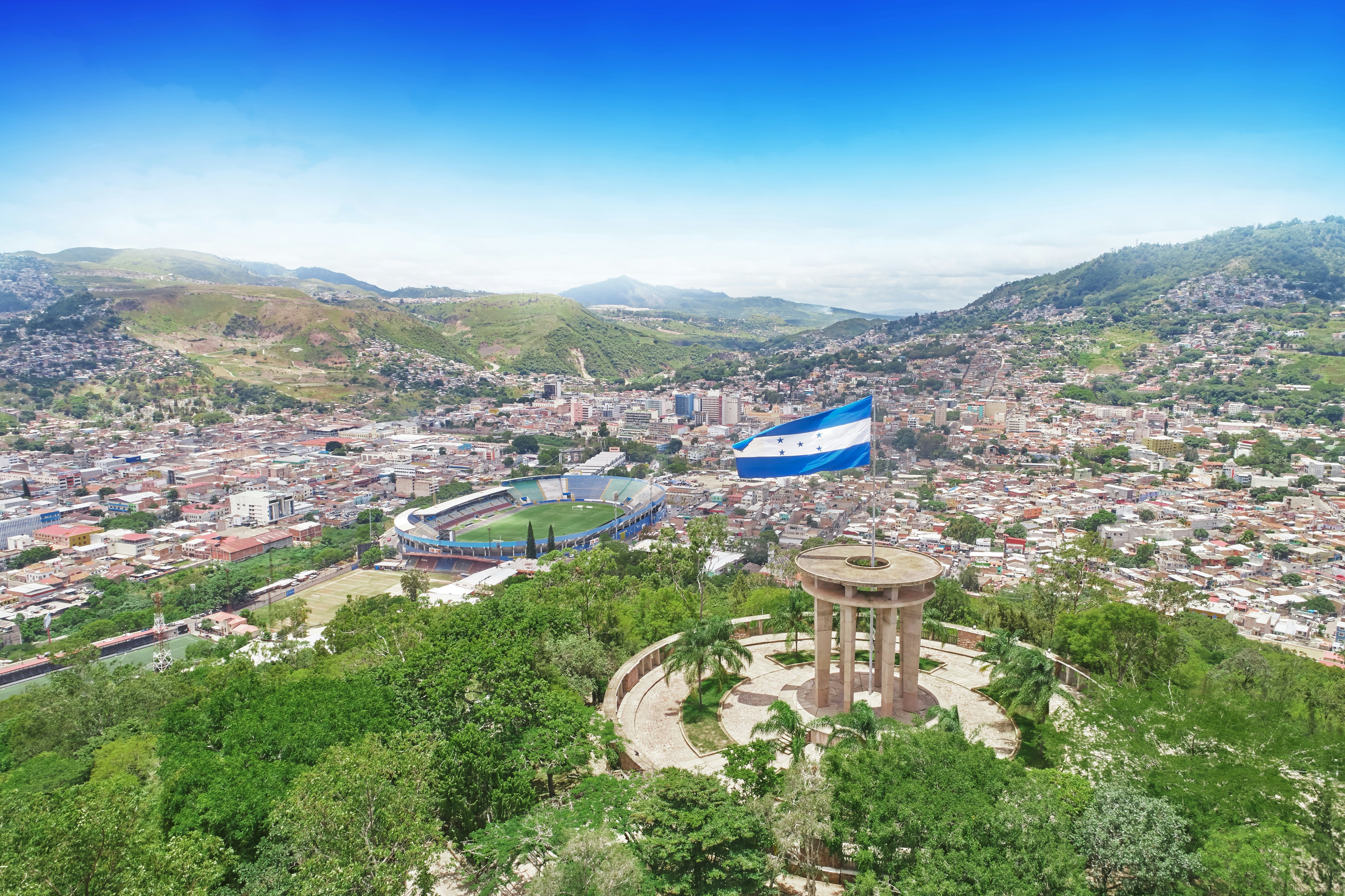 A city in a hilly landscape. A blue and white flag flies from a hilltop and a large sports stadium stands among smaller buildings in the valley below.