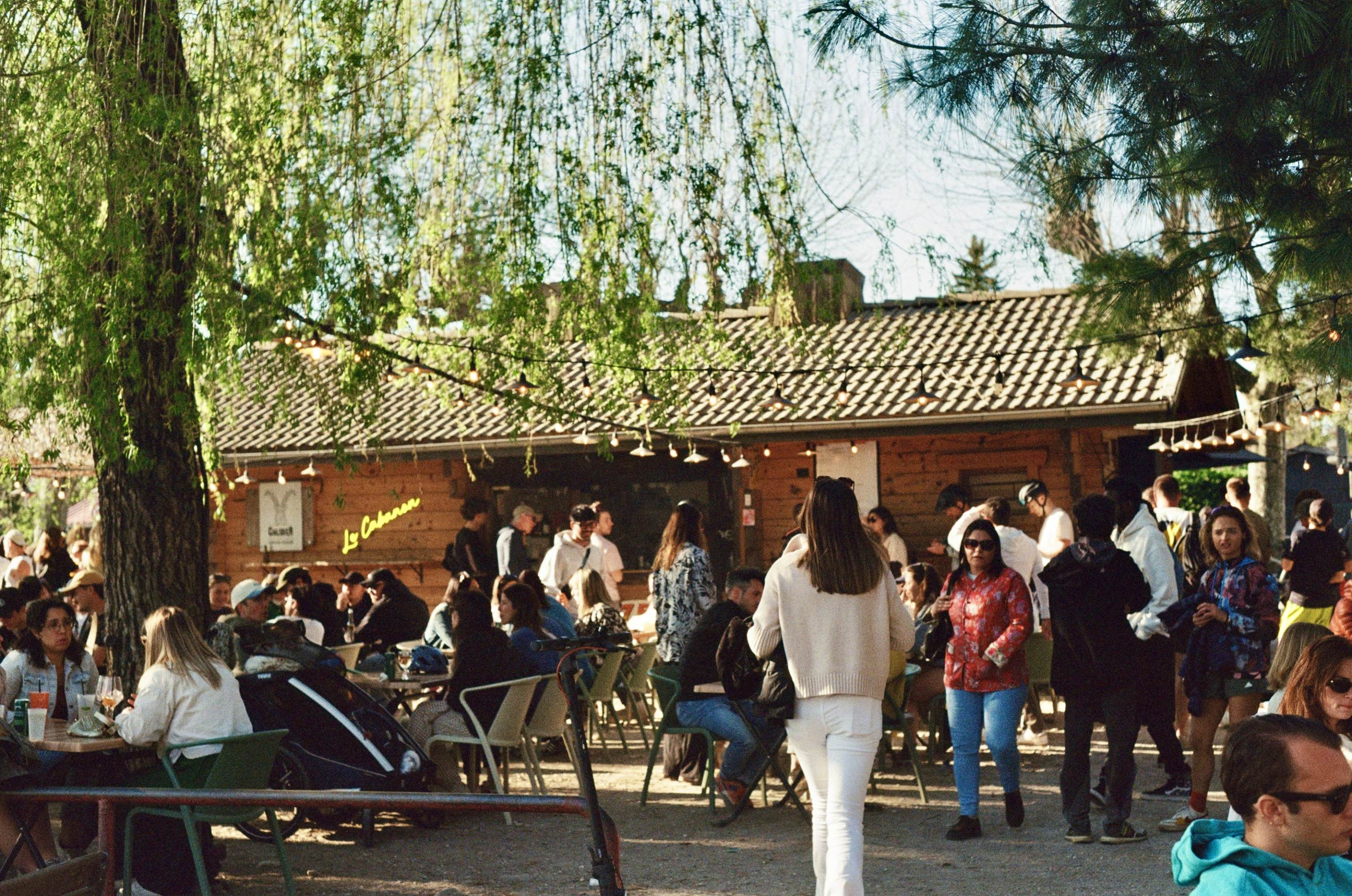 People at and around picnic tables outside at a bar in France; there is a large tree providing shade, and a wood building is in the background.