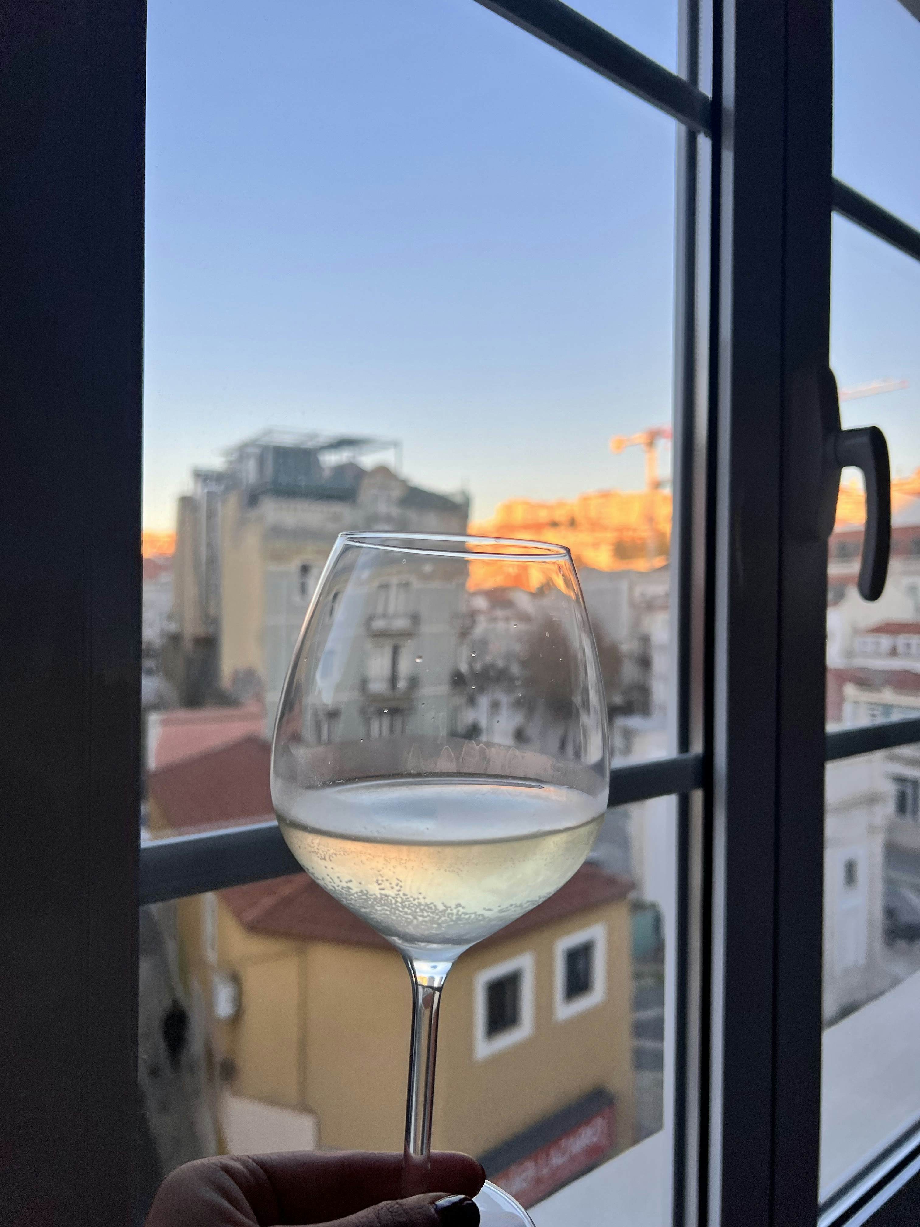A clear glass of white wine in front of a window looking out at a city at sunset.