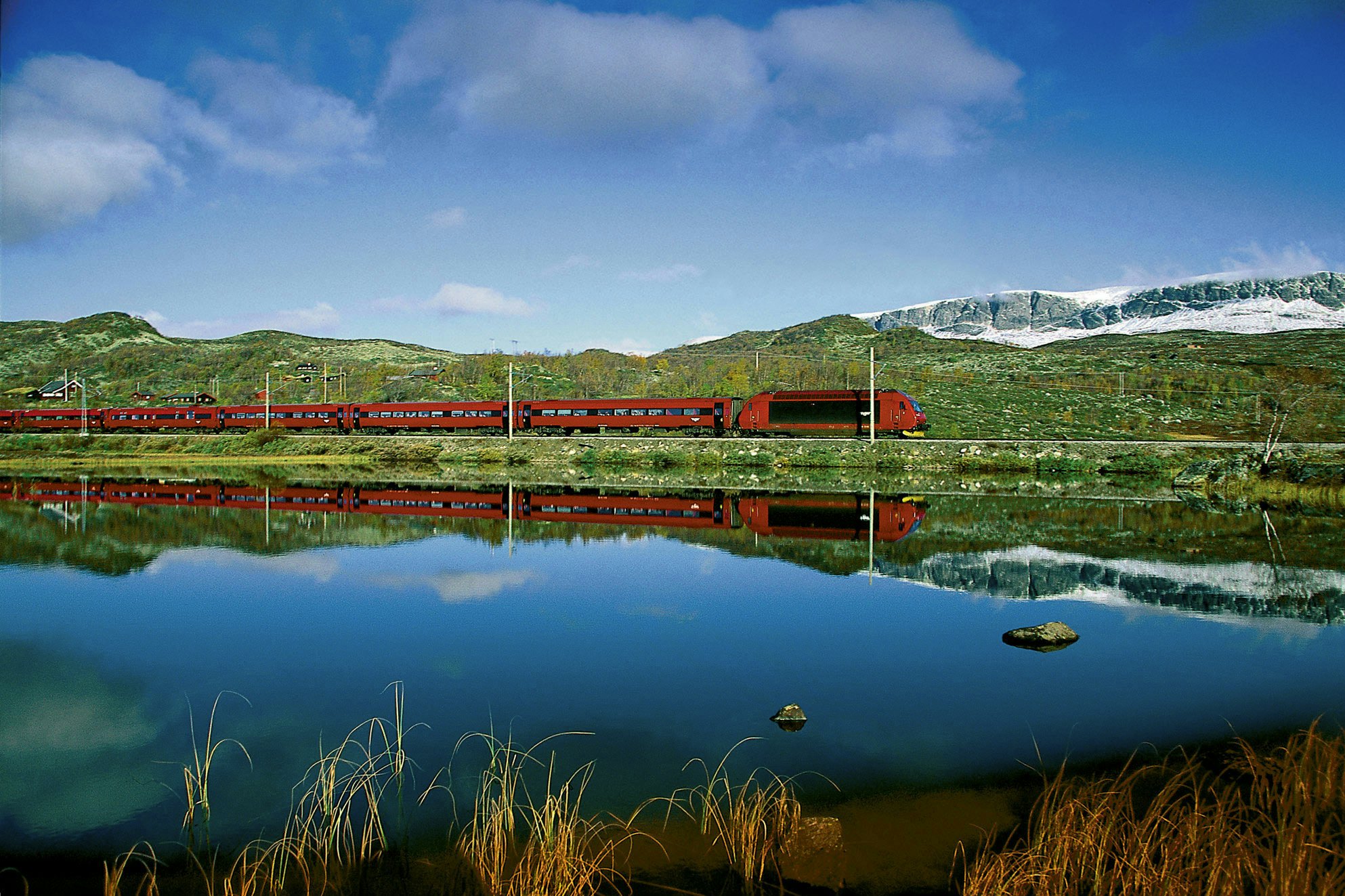 A red train is reflected in the calm surface of a blue lake.