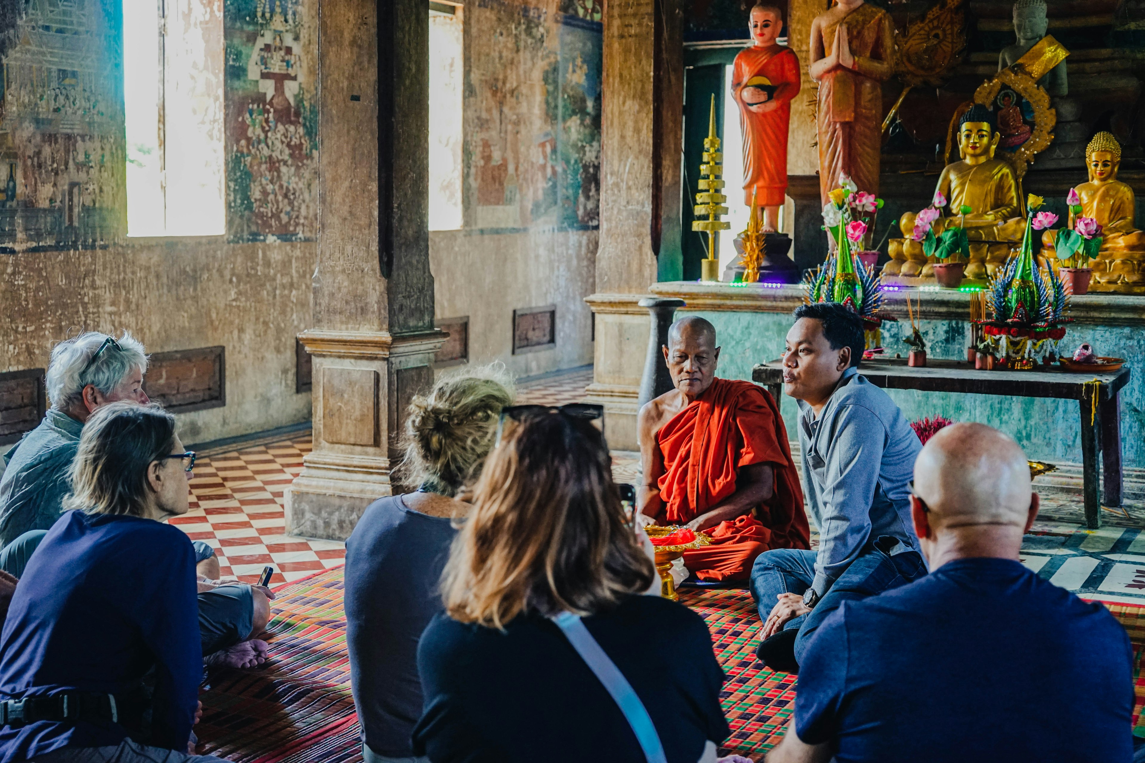 A group of tourists sit on a rug in front of a Buddhist monk and interpreter.