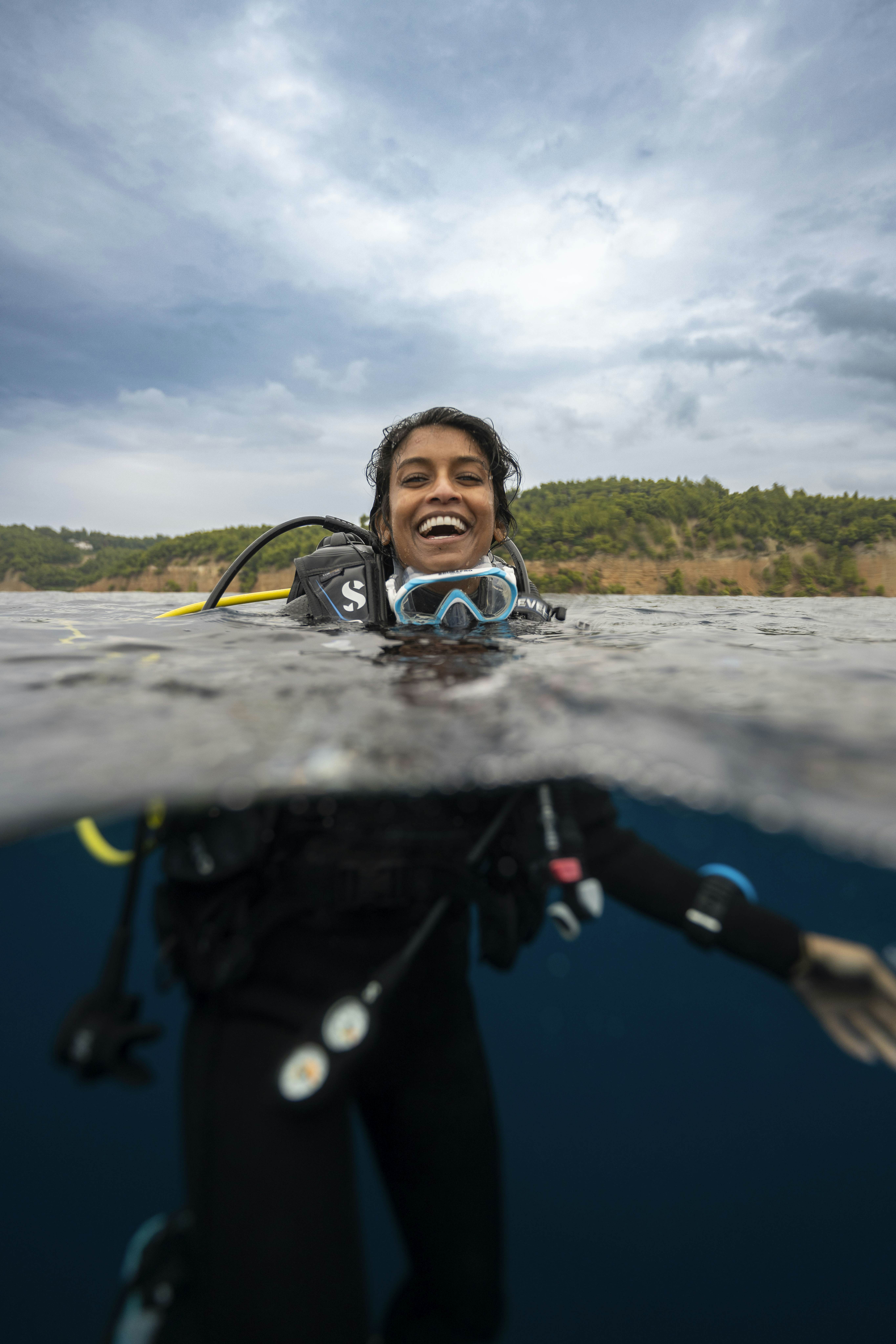 A woman smiles with her head above the water; she has a scuba mask below her chin, and her wetsuit if visible below the water line. 
