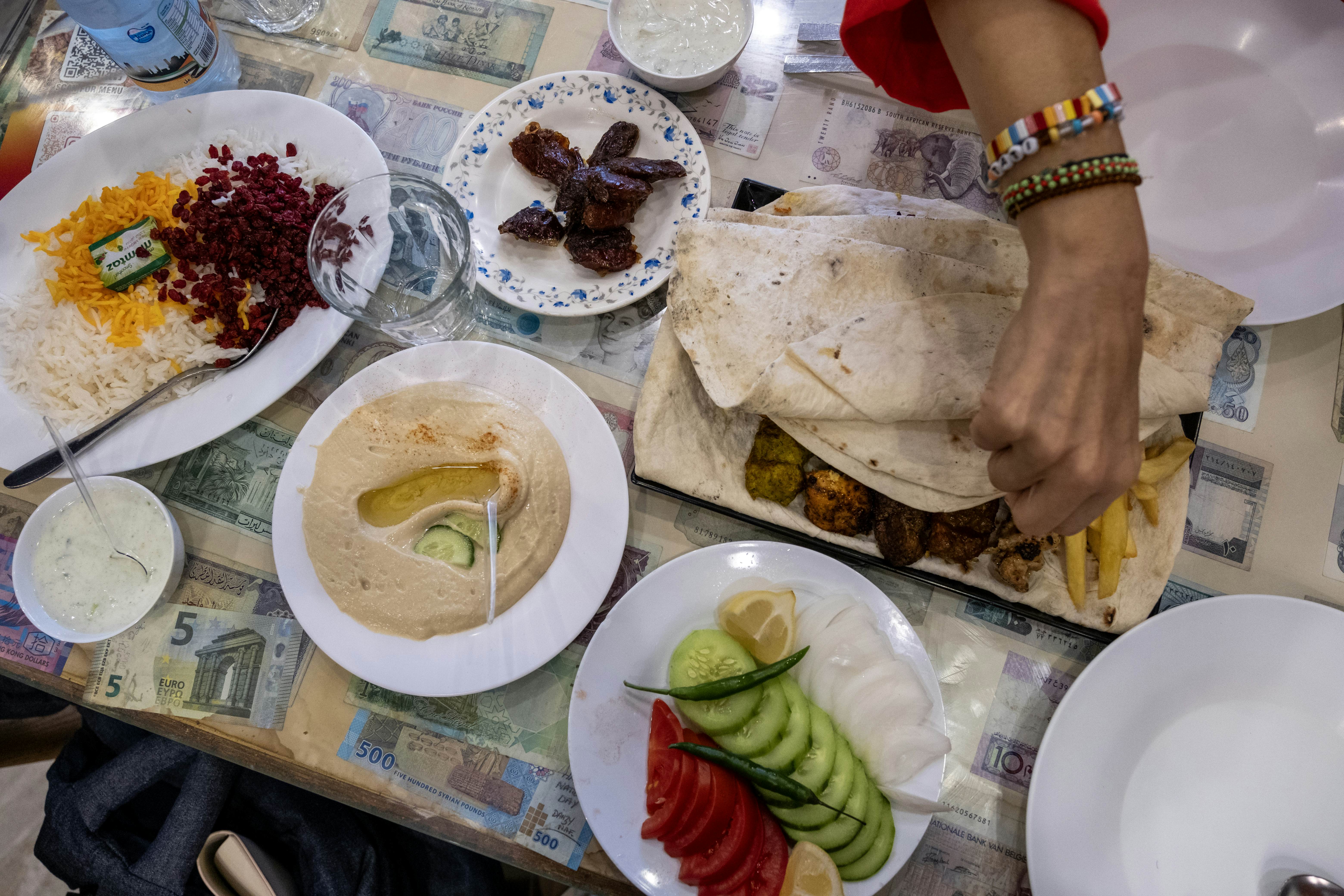 A plate of cucumbers and tomatoes sits alongside a plate of hummus and a kebab in pita bread