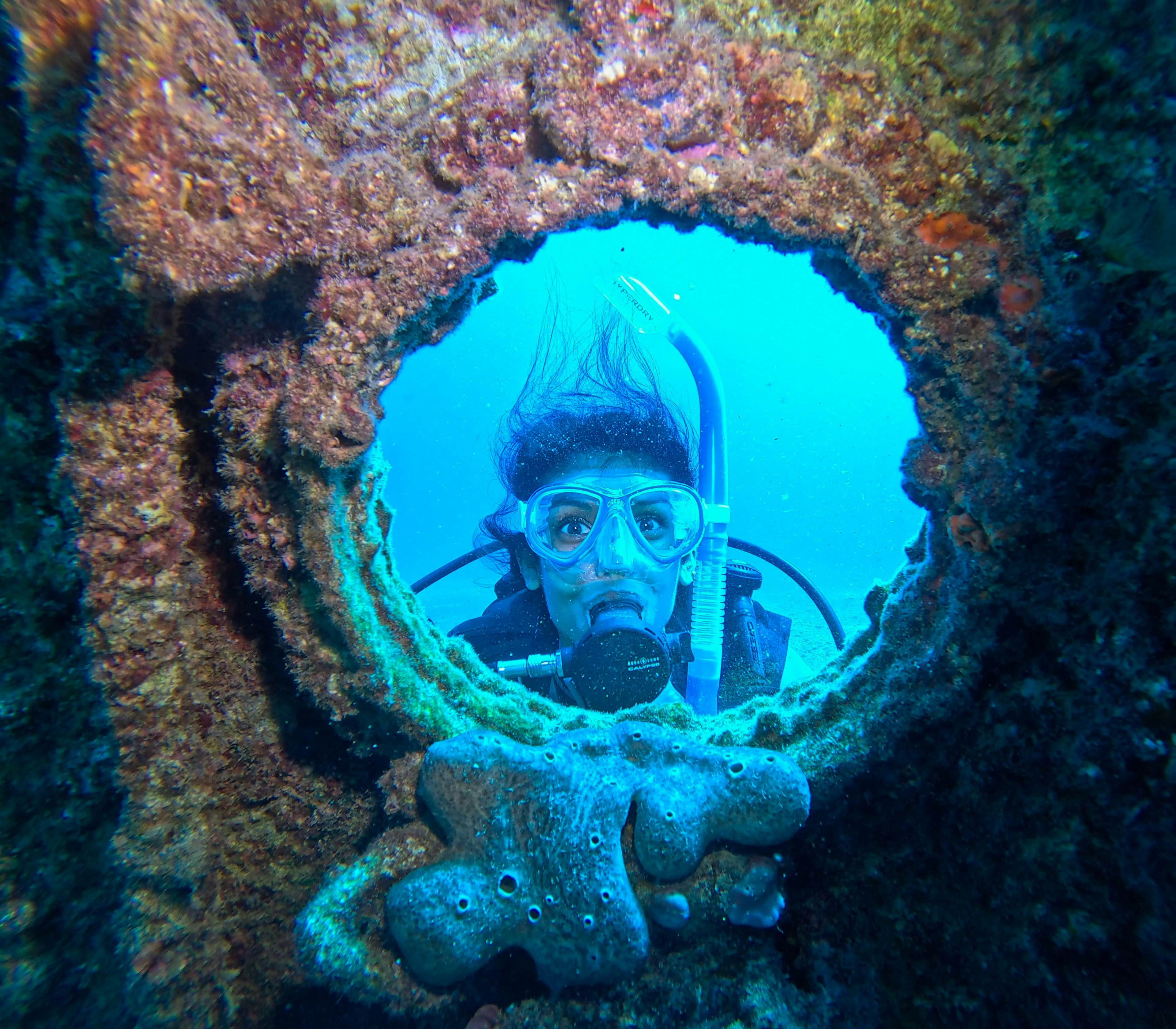 A woman in a scuba mask underwater peers through a porthole.
