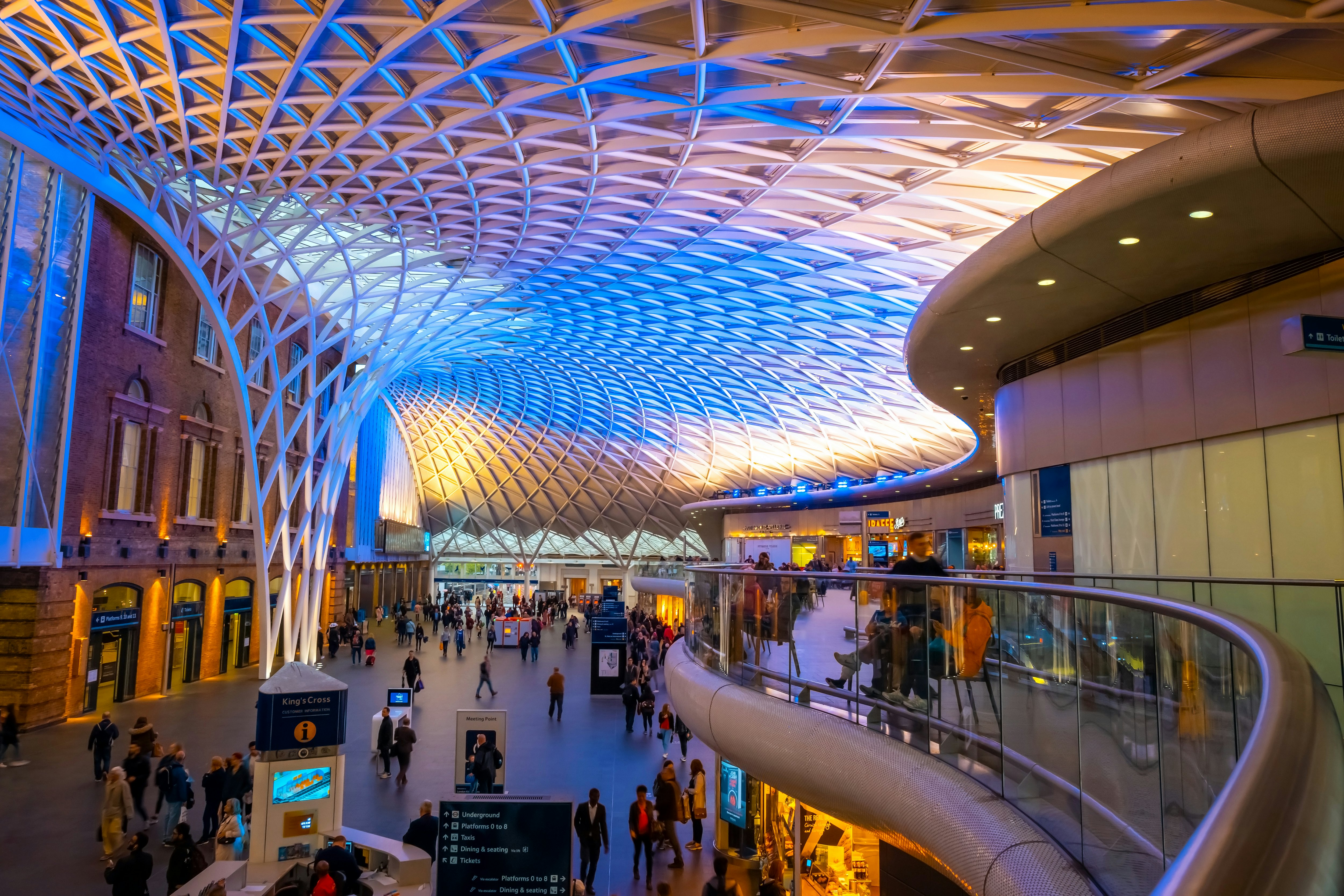 People pass through a station concourse with a mezzanine balcony and a curved roof.