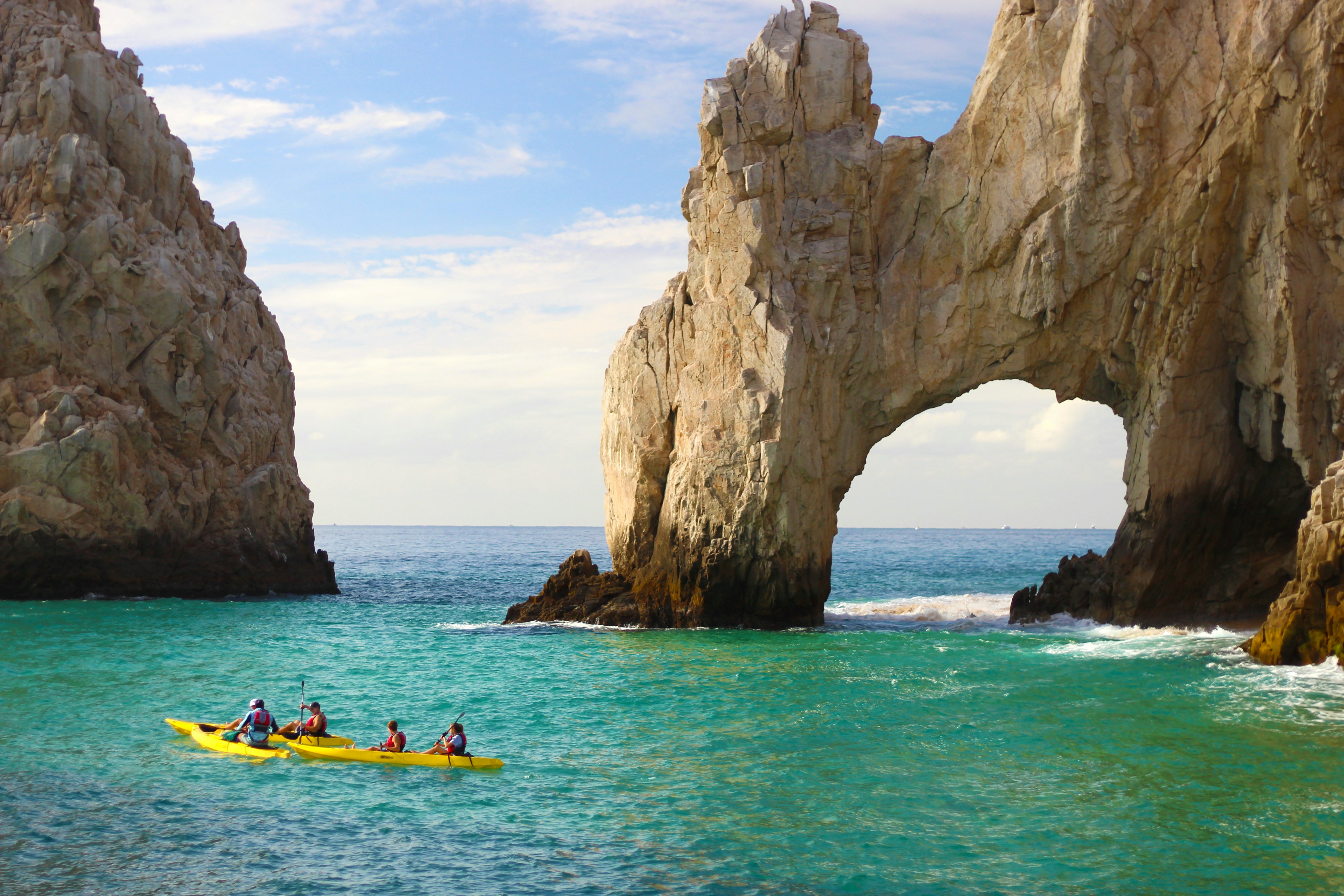 People in yellow kayaks paddle toward a rock arch formation in the sea.