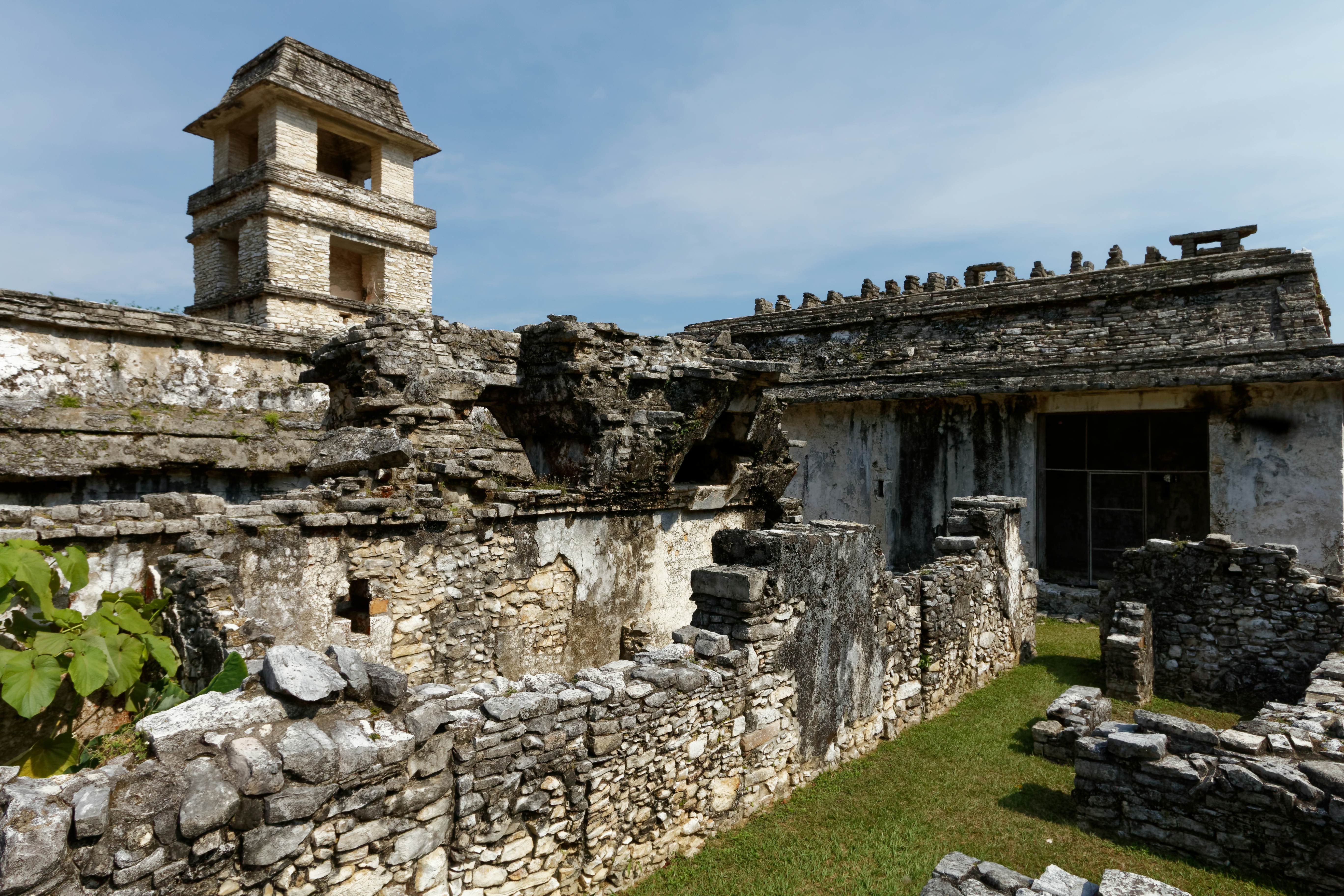Ancient stone ruins of a low building with a tower in the background on a sunny day.