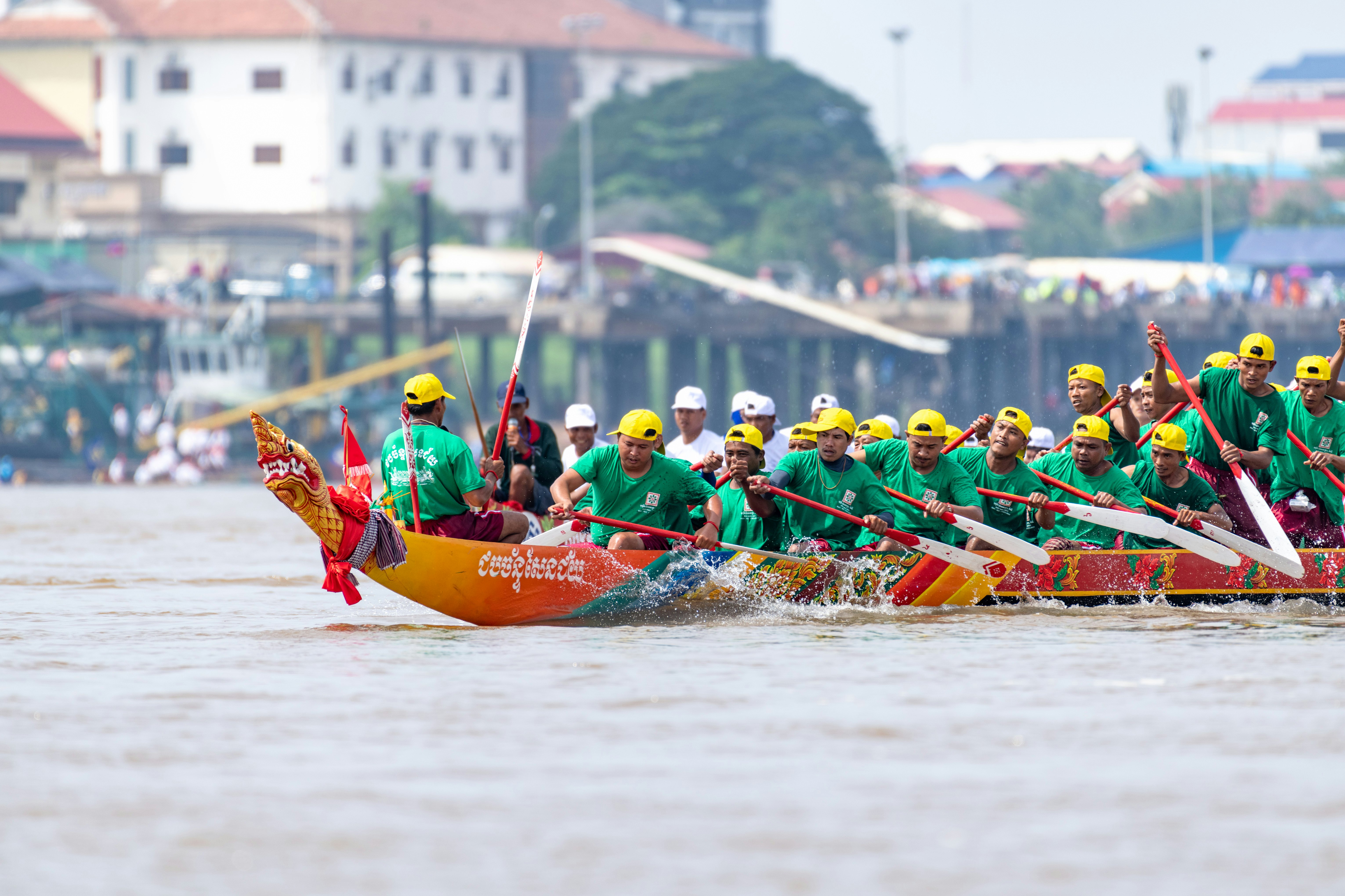 A group of racers paddling in a long boat. They are wearing green shirts and yellow baseball caps.